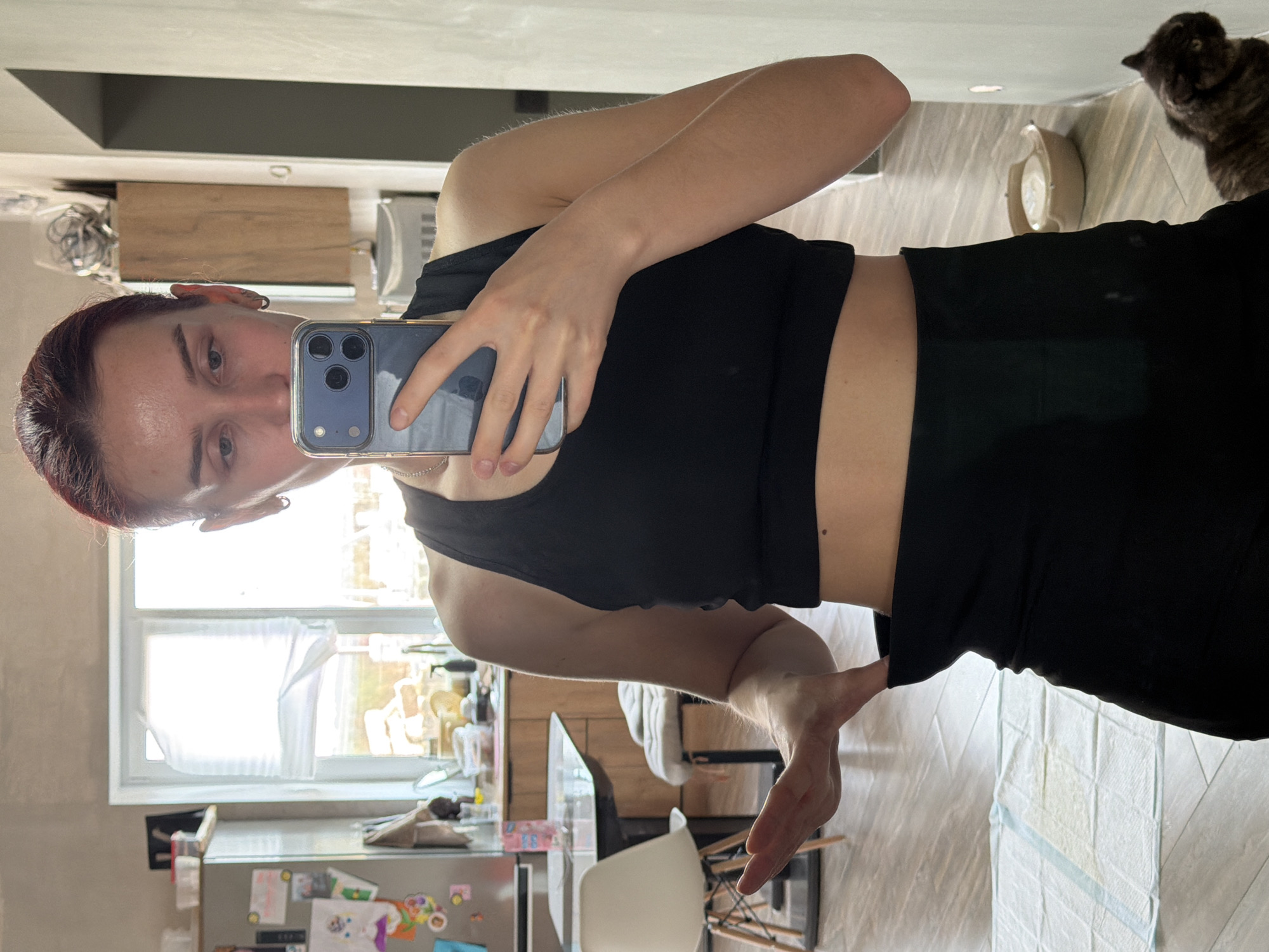 A person is taking a selfie with a phone, wearing a black tank top and black pants. The person has short, reddish-brown hair and is positioned in a kitchen or dining area. A window is visible in the background, allowing daylight into the room. Kitchen appliances and objects are scattered around the room. A cat is visible in the upper right corner of the frame. The location is the city of Chaikovsky, Russia.