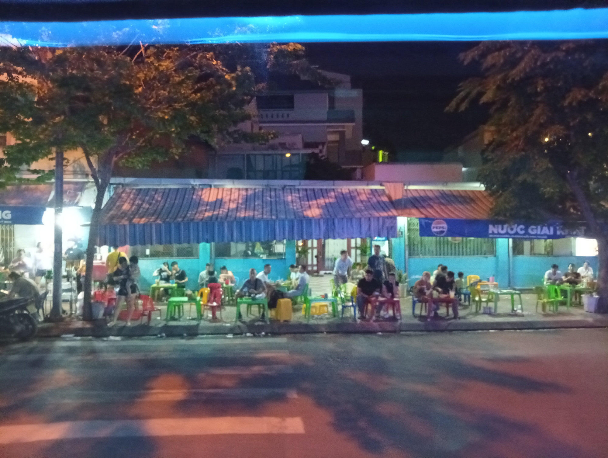 The image depicts a street-level view of an outdoor seating area at night in Nha Trang, Vietnam, likely a restaurant or cafe. A bright blue awning stretches across the width of the establishment, shading several tables and chairs where patrons are seated. The seating area is open to the street, with a sidewalk and road visible in the foreground.
People are scattered throughout the seating area, engaged in various activities. Some are seated at tables, seemingly dining or socializing. Others appear to be standing or walking, moving between tables or entering/exiting the establishment. The lighting is dim, typical of nighttime, with artificial lights illuminating the area and casting shadows. Trees with foliage are visible to the left and right of the establishment, suggesting it is located on a tree-lined street.
A sign with the Pepsi logo and Vietnamese text that translates to "Soft Drinks" is visible on the blue awning. A motorcycle is parked on the left side of the image, near the entrance to the seating area. The overall impression is a lively, casual nighttime scene in a Vietnamese city.