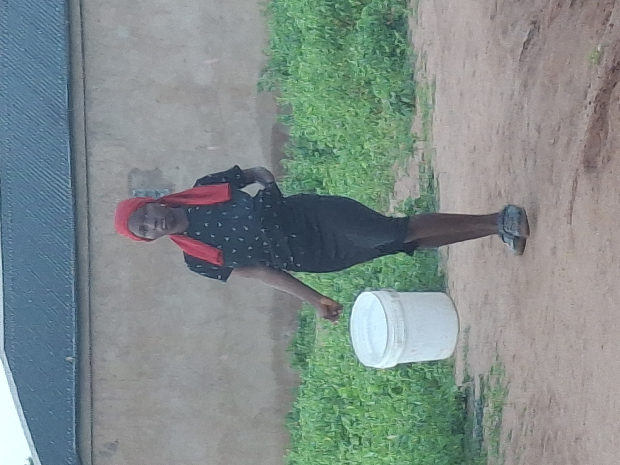 In Tumfure, Nigeria, a young person with dark skin and a red headscarf stands outdoors, holding a white bucket filled with water. The individual is wearing a black, short-sleeved shirt with a small pattern and a black skirt. The person is looking towards the camera with a neutral expression and has their left hand near the bucket. The ground in front of them is dirt, and behind them is a wall and lush green foliage. The sky is overcast.
