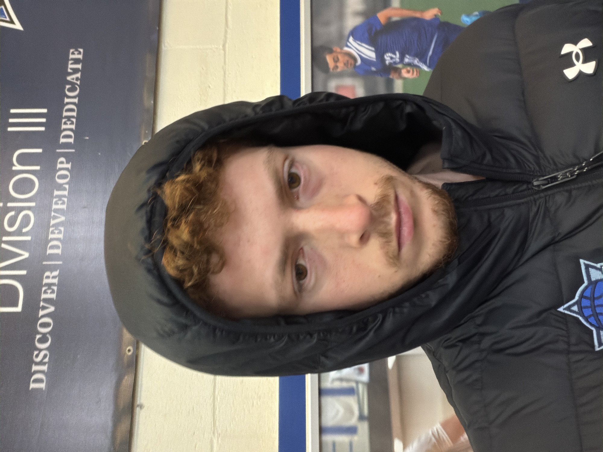 A man with red hair and a beard is wearing a black hooded jacket. He is indoors. Behind him are walls and signs, one of which says "Division III" and another which shows a soccer player. The location is the city of New York, United States of America.