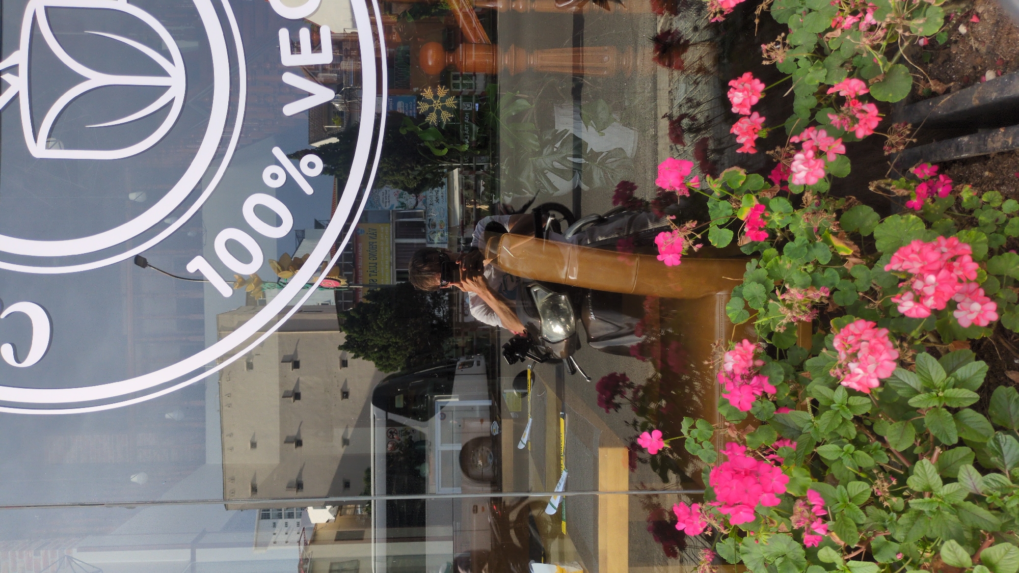The image shows a reflection in a storefront window in Da Lat, Vietnam. In the reflection, a person is taking a selfie with a smartphone while sitting on a scooter. The person is wearing a white t-shirt and sunglasses. Behind the person, the reflection shows buildings, trees, and a bus. In the foreground of the reflection, there are pink flowers. The storefront window has a circular logo with "100% VEG" written on it.