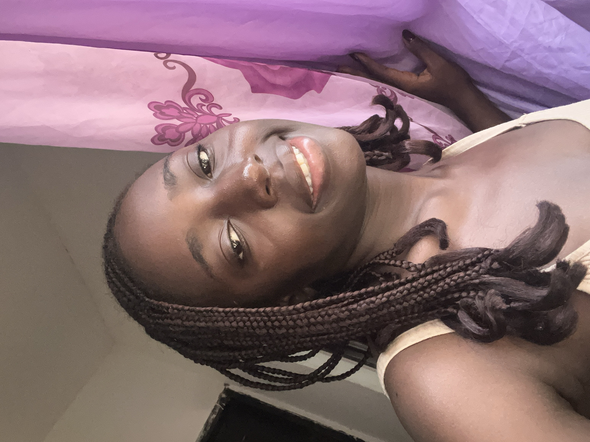 A close-up, upward-angled portrait features a young Black woman lying down, looking directly at the camera with a broad, joyful smile. Her head is positioned to the left, with her body extending towards the right side of the frame. She has long, dark box braids, some of which are styled around her head and shoulder. Her skin appears radiant, with a subtle sheen. She is wearing a light-colored (possibly cream or light yellow) tank top or camisole.
The background behind her head and upper body consists of two fabric layers: a light pink or lavender fabric with a darker pink, ornate floral pattern, and a solid, darker purple fabric further behind. One hand is visible near the top right, resting on the purple fabric. Below her, a plain, light brown surface is visible. The lighting appears soft and natural, highlighting her features. Her expression conveys happiness and comfort. No visible text is present. The scene is set indoors in Abuja, Nigeria.