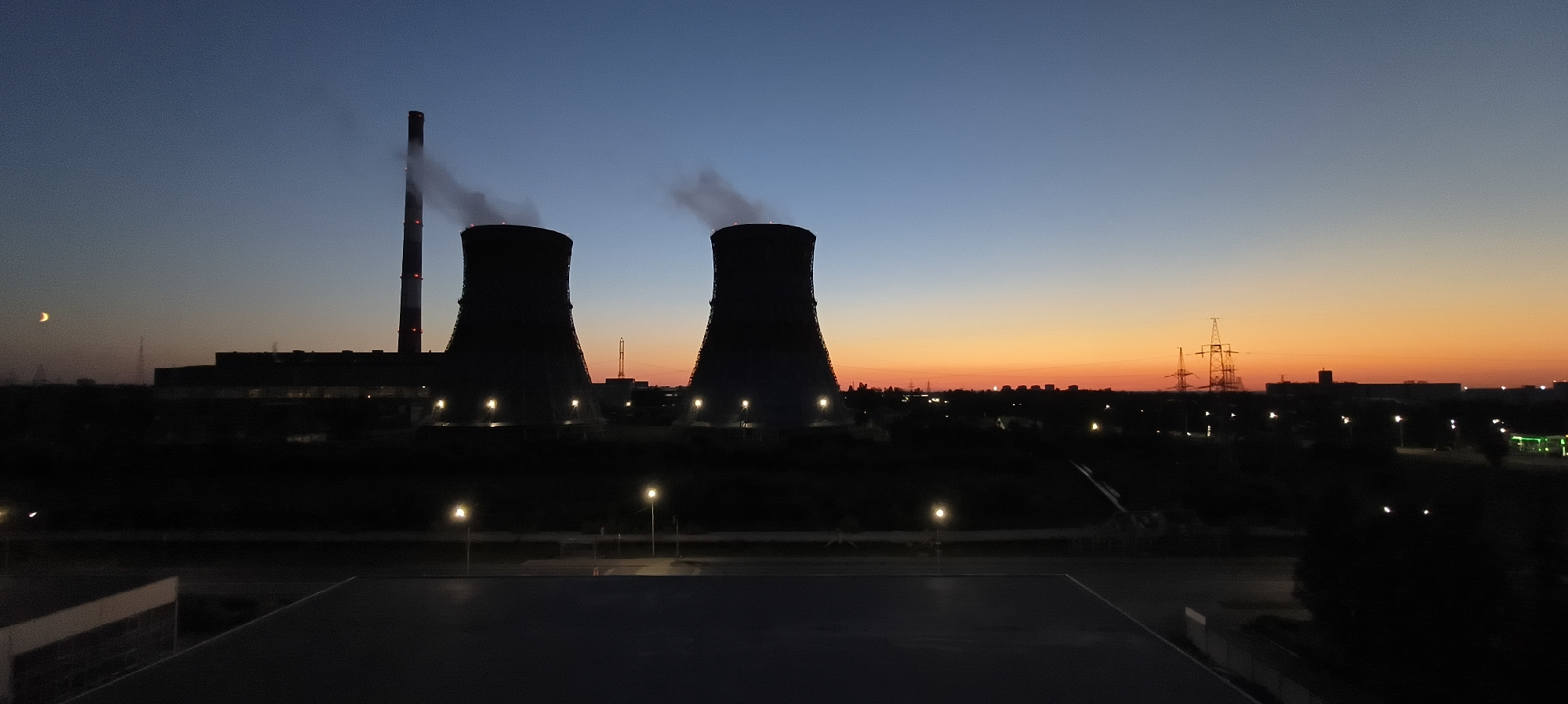 This is a wide-angle, low-angle shot of a power plant at dusk. In the foreground, there is a dark, flat surface, possibly a roof, with a light colored stripe on the left side. Beyond this, there is a dark landscape with streetlights and trees. In the middle ground, two large, dark, cooling towers are prominent, with smoke or steam emitting from their tops. A tall smokestack with red and white bands is to the left of the cooling towers, also emitting smoke. The background features a dark industrial complex, power lines, and buildings. The sky transitions from a deep blue at the top to a vibrant orange and yellow near the horizon, indicating sunset or sunrise. A sliver of a crescent moon is visible in the upper left portion of the sky. The overall scene is dark and industrial, illuminated by artificial lights and the fading sky.