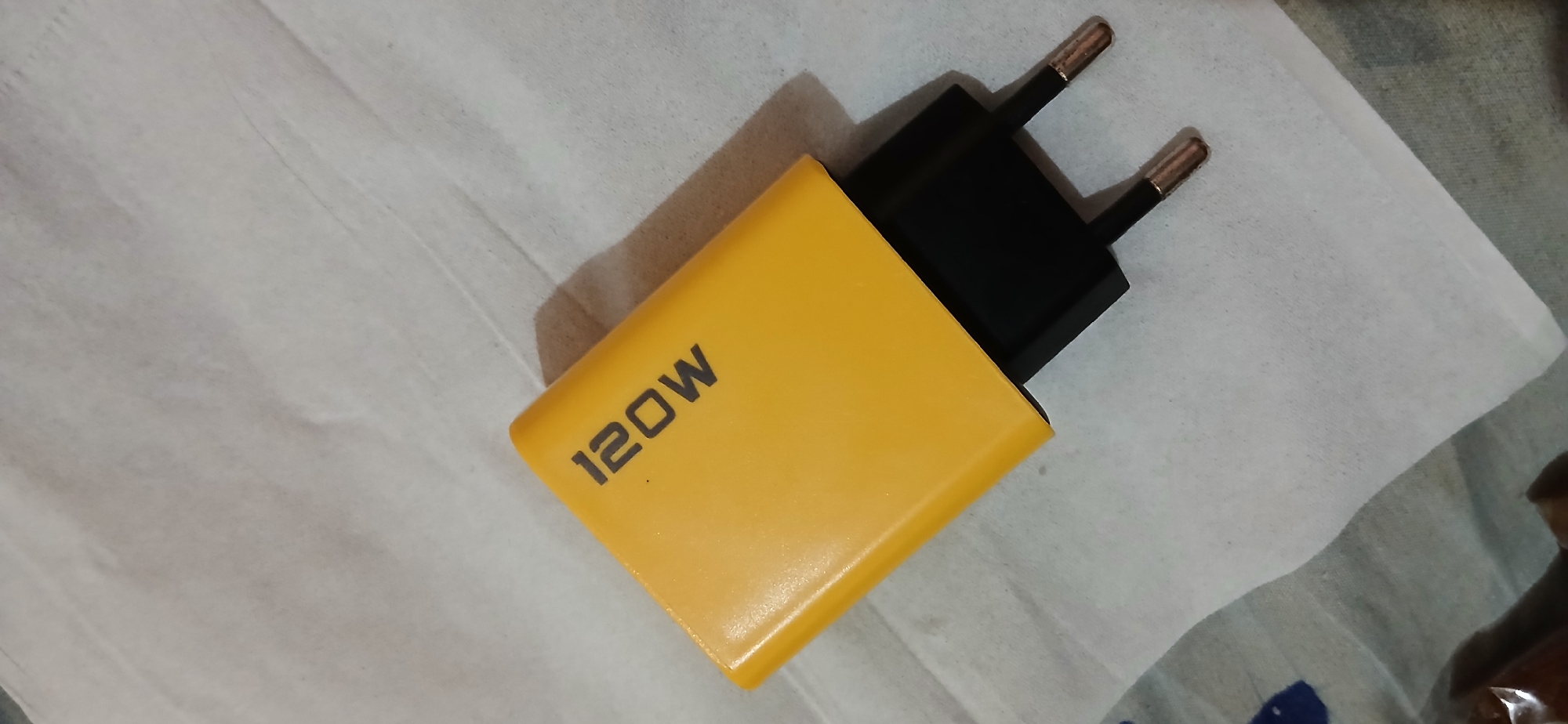A yellow charger with "120W" printed in black is lying on a white surface. The charger has a black plug with two prongs. The background is a textured white cloth. The image was taken indoors. There is no information about the location in Samarinda, Indonesia.