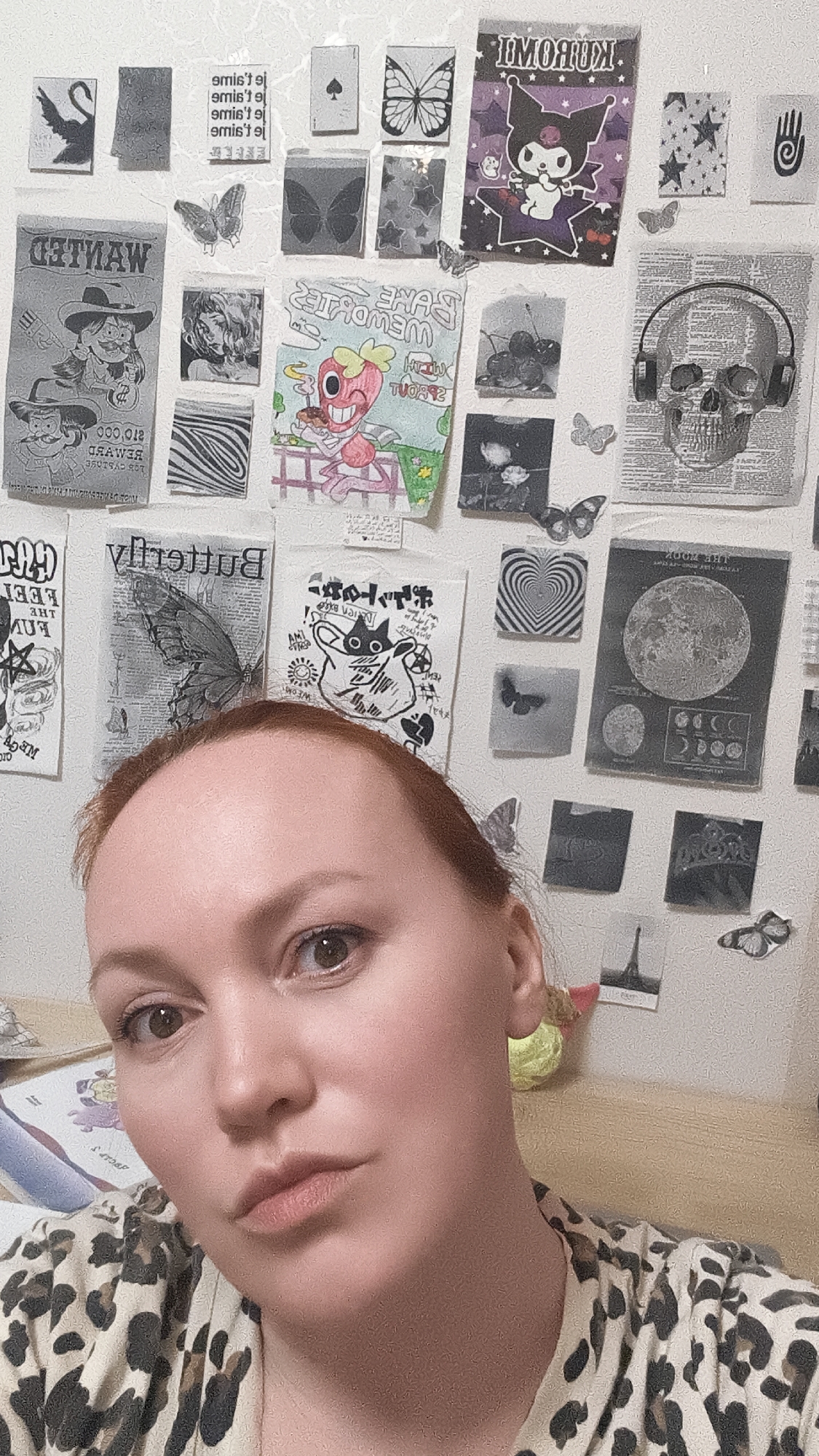 A close-up, indoor shot features a woman with light skin, reddish hair, and brown eyes. She is wearing a leopard print top. The background is a white wall covered in various printed images and stickers. These include a "Wanted" poster, a "Kuromi" poster, a skull with headphones, butterflies, a moon phases chart, and several other miscellaneous graphics. The environment suggests a personal space, possibly a bedroom or study area. A portion of a desk or table is visible in the foreground.