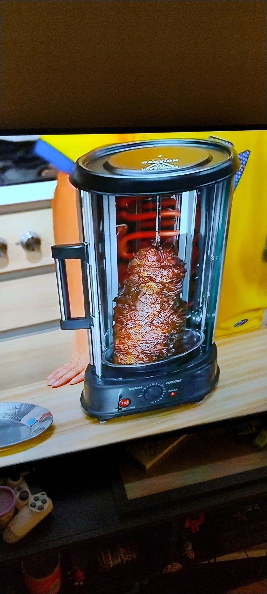 A vertical, close-up shot shows a rotisserie cooker with a large piece of meat cooking on a spit. The cooker is black and clear plastic, with red heating elements visible inside. The meat is a deep brown color, indicating it is well-cooked. The cooker is placed on a light-colored wooden surface. To the left of the cooker, a hand with pale skin is visible, resting on the surface. A person wearing a bright yellow shirt is partially visible in the background on the right. The lighting appears to be from a television screen or monitor, casting a slightly diffused glow on the scene. The overall environment suggests an indoor setting, possibly a kitchen or living area. There is no specific information in the image to determine its location in Ishøj, Denmark.