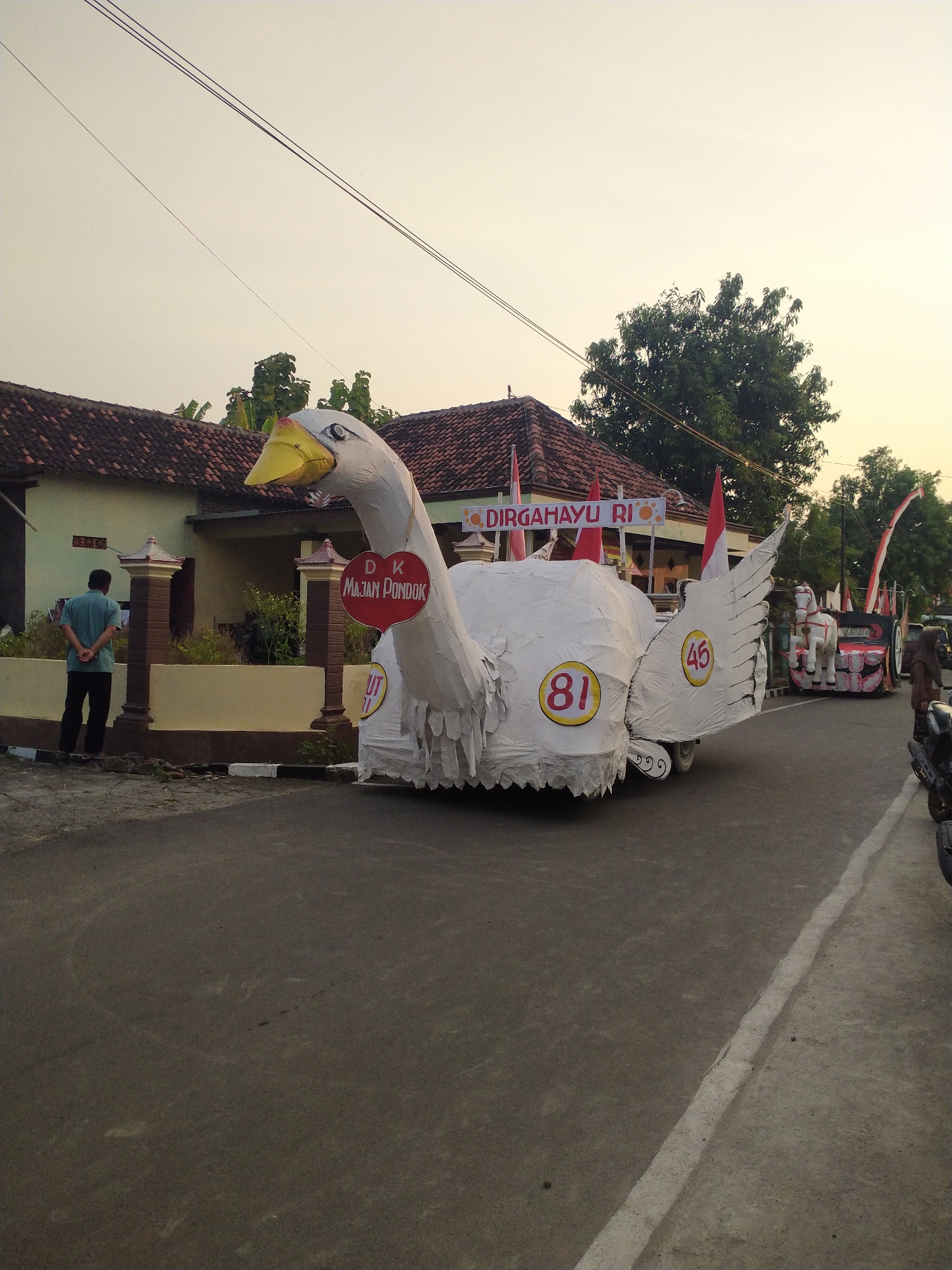 In the city of Pondok, Indonesia, a street is lined with houses and trees under a clear sky. A parade float, designed as a large white swan with a yellow beak, dominates the foreground. The float is decorated with red and white flags and displays the text "DIRGAHAYU RI" and "DK MAJAM PONDOK". Two circular emblems are visible on the float's body, one with the number "81" and another with "45". Behind the swan float, another vehicle in the parade features a white horse figure on a red base. A man in a blue shirt and black pants stands by the side of the road, facing away from the camera. A portion of a motorcycle is visible in the right side of the frame. The scene appears to be a local celebration or event.