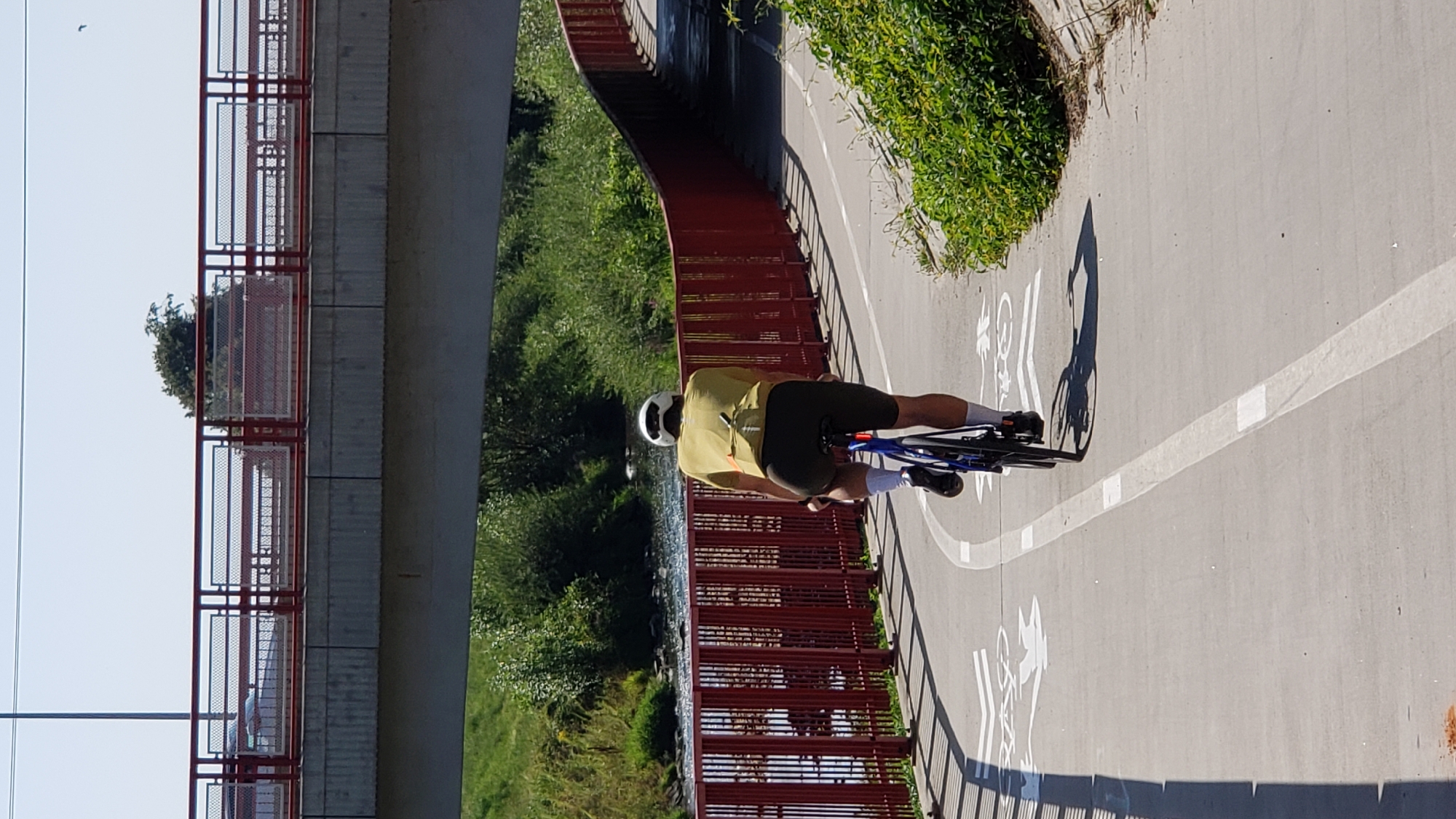 A lone male cyclist wearing a yellow t-shirt, dark shorts, and a white helmet is cycling on a paved path. He is viewed from behind, and his bicycle is blue. The path is marked with white lines and bicycle symbols. The path runs alongside a red-railed embankment with a river visible on the other side. A concrete bridge with red railings spans the scene above, and the sky is clear and blue. The location is the cycling path along the Torysa river in Prešov, Slovakia.