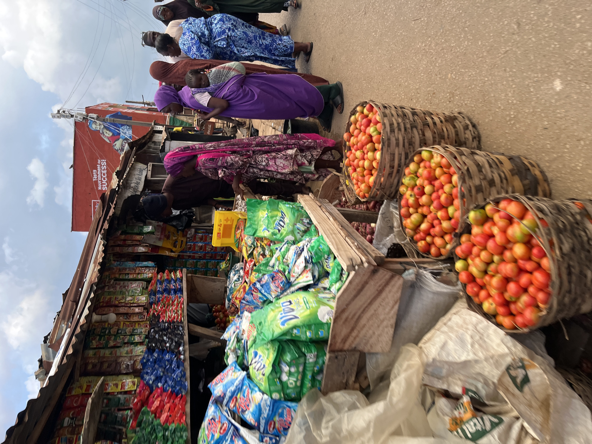 This image captures a bustling outdoor market scene in Jos, Nigeria, during the daytime under a cloudy sky. The scene is likely a street market with informal stalls and a mix of vendors and shoppers.
In the foreground and mid-ground, several women are present. Most are dressed in modest, traditional attire, including head coverings. One woman in a vibrant purple dress is seen carrying a child on her back, secured with a fabric wrap. Another woman wears a blue patterned dress. The women appear to be moving through or standing within the market space, engaging in daily activities.
To the right, three large, round woven baskets are prominently filled with fresh, ripe tomatoes, exhibiting a mix of red and greenish-orange hues, likely awaiting purchase. On the left side of the frame, an open-front stall or shop displays a wide variety of goods. Its roof is made of corrugated metal sheets. Inside, numerous packaged items are visible, including large green bags of "Vim" detergent and smaller blue bags, possibly another detergent brand like Omo. Below these, shelves are stacked with rows of smaller, colorful packets, possibly spices, snacks, or condiments. A "Coca-Cola" logo is faintly visible on an item near the purple-clad woman.
In the background, further down the street, a large red billboard features an image of a woman and child, with the visible text "Tasty Nourishment for SUCCESS!" Utility lines crisscross against the sky near the billboard. A white sack near the tomato baskets has "VITAL" partially visible. The overall atmosphere suggests a lively and typical market environment in an urban setting.