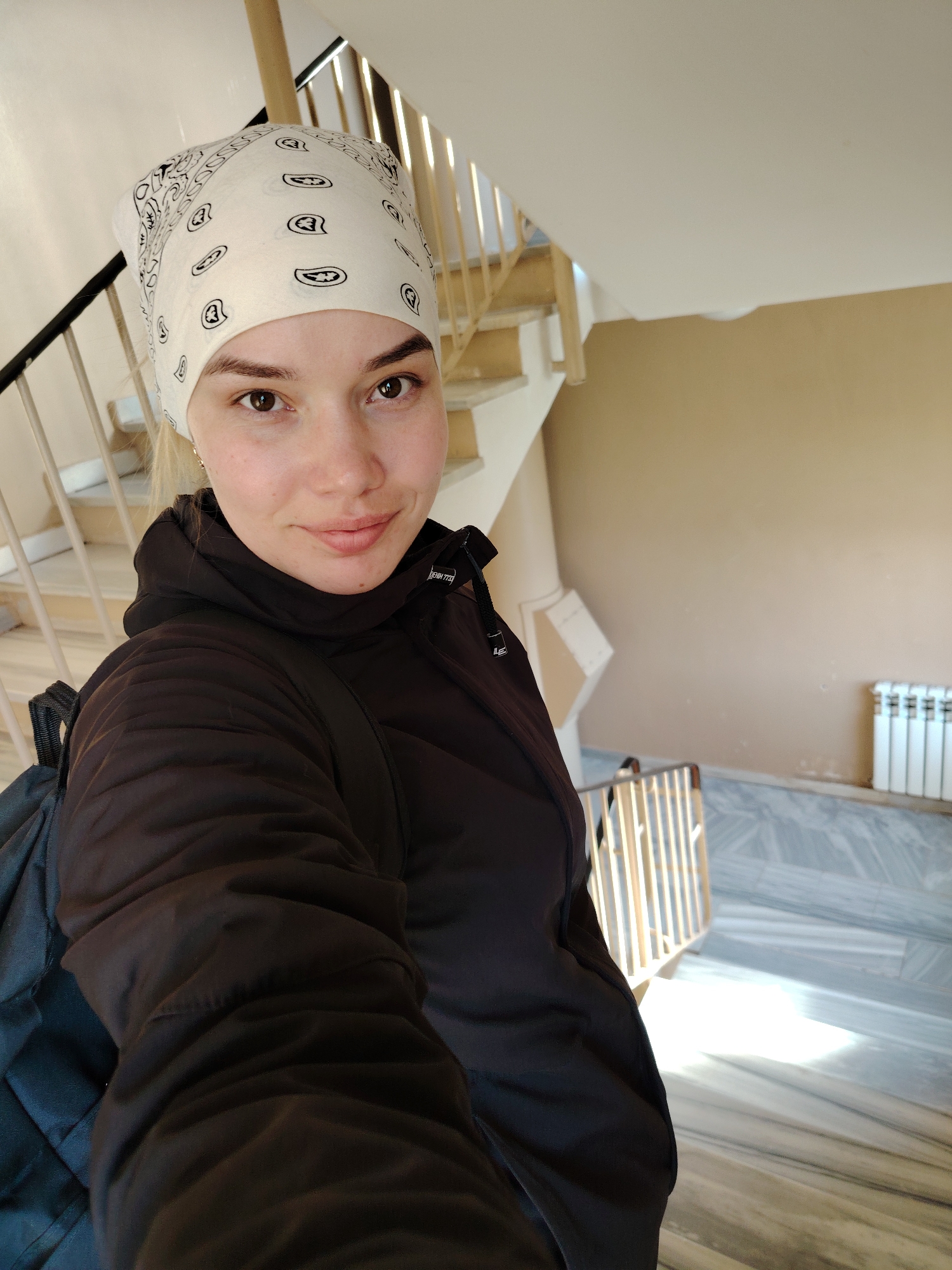 A woman stands in what appears to be the stairwell of an interior space. She wears a dark, puffy jacket, a light-colored bandana with a patterned design, and a backpack. The stairwell features wooden stairs with black metal railings and a radiator against a beige wall, all situated on a marble-like floor. The location may be in Markovskii, Russia.