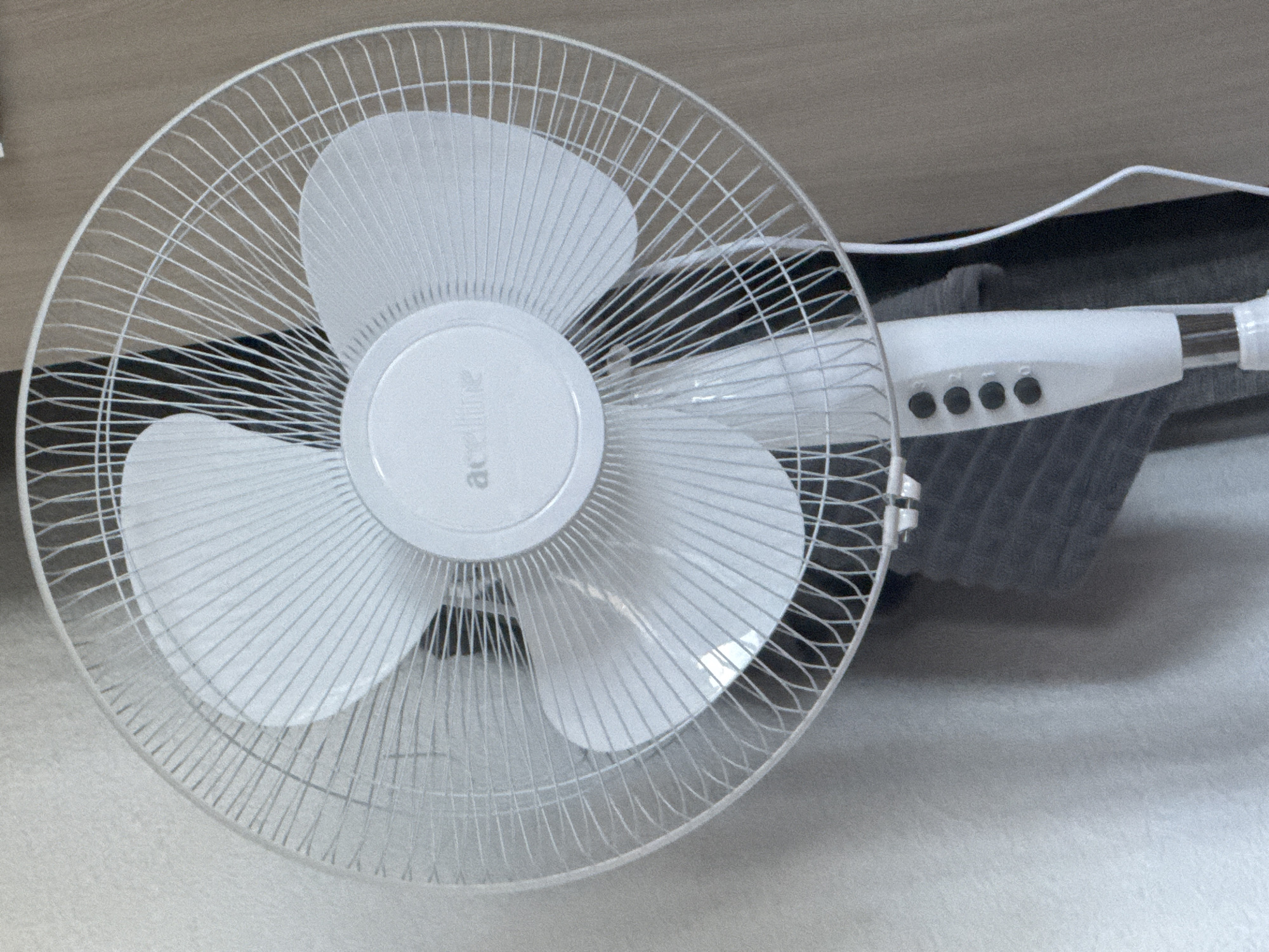 An upright white fan with a circular guard and three white blades is positioned against a lightly textured, pale wall. A dark garment is draped behind the fan, and a power cord extends from the base. The fan's central housing displays a white logo with faint text. The base of the fan, also white, features four black control buttons labeled "1", "2", "3", and "4". The scene appears to be indoors, possibly in a residential setting in the city of Krasnye Baki, Russia.