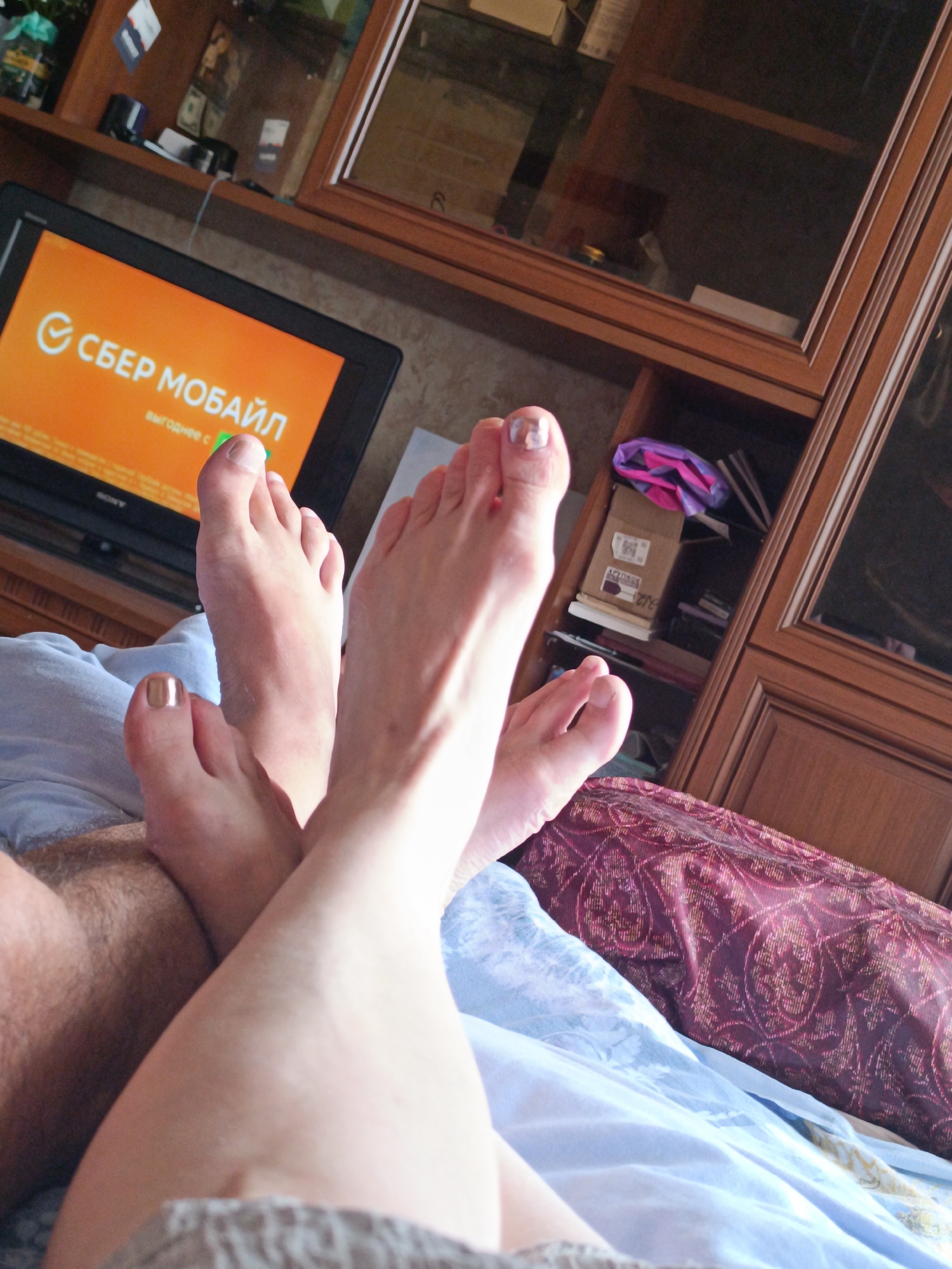 The image shows a close-up view of two pairs of feet, one with metallic gold nail polish and the other with a marble-patterned nail polish. The feet are resting on light blue and burgundy patterned fabric, likely bedding. In the background, a television screen displays an orange screen with white text that appears to be a logo or advertisement. Behind the television, a wooden cabinet with glass doors is visible, containing various items, including what appear to be books or boxes. The lighting suggests an indoor environment.