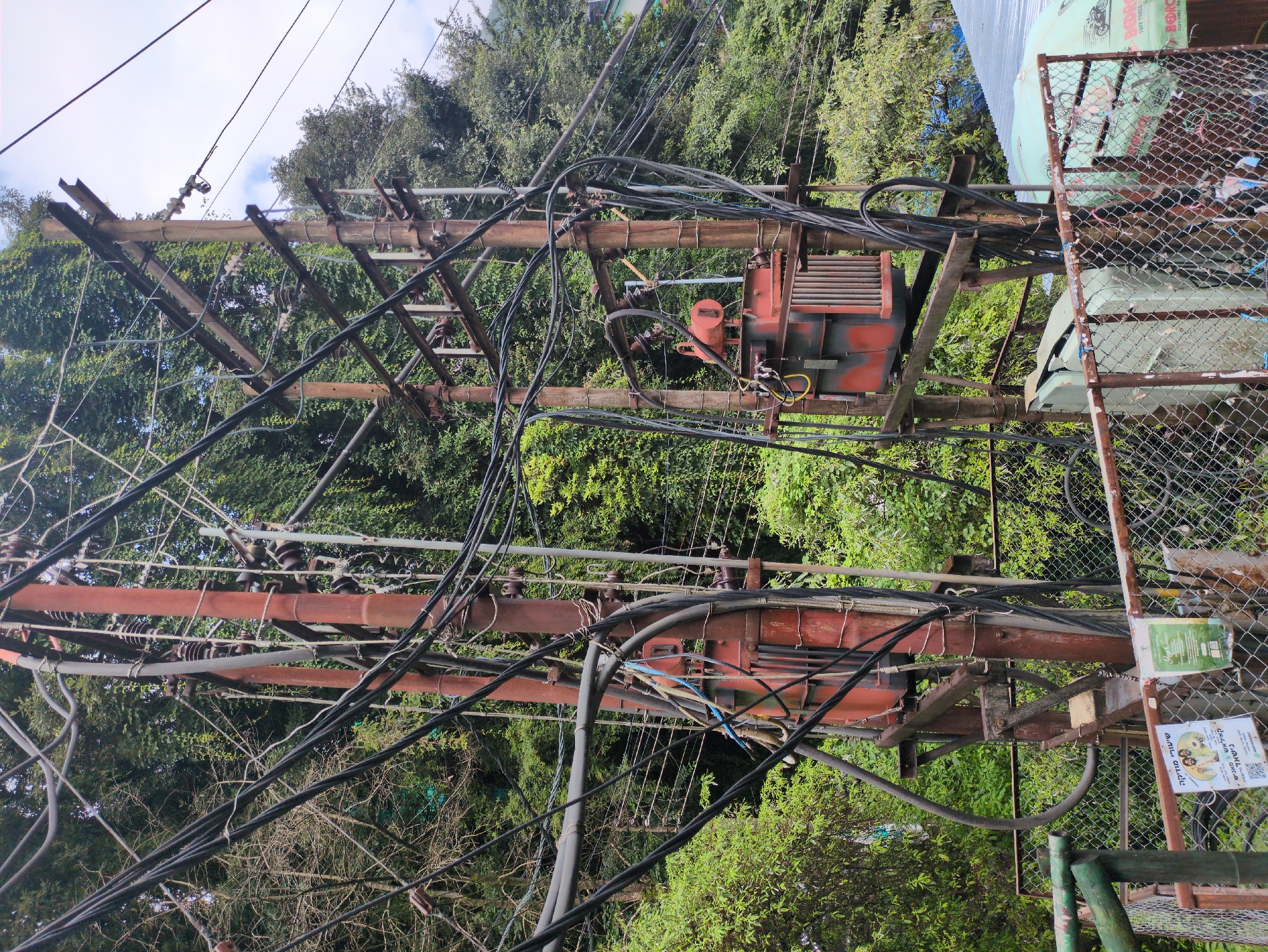 In the city of Bhagsu, India, a dense tangle of overhead electrical wires dominates the scene, suspended between two weathered, rust-colored utility poles. These poles, constructed of what appears to be metal, are adorned with numerous ceramic insulators, supporting the complex network of black cables.
Mounted on the right-hand pole, a large, red and grey electrical transformer hangs precariously. Its metallic casing shows signs of age, with vertical ribbed panels on its lower section. Below the transformer, another, smaller grey electrical box is visible.
The background is a lush tapestry of dense green foliage, indicative of the natural surroundings of Bhagsu. The sky above is overcast with a few patches of blue peeking through. In the foreground, a rusty metal fence with chain-link mesh encloses the area. Two posters are attached to this fence. The poster on the left features images of two men and text in Hebrew. The poster on the right has text in English and some green graphic elements. Further to the right, a pale blue corrugated metal roof is partially visible, along with a green umbrella with the text "BEDROCK TUFF TYRES" printed on it. The overall impression is one of functional utility integrated into a verdant, possibly hilly, urban environment.