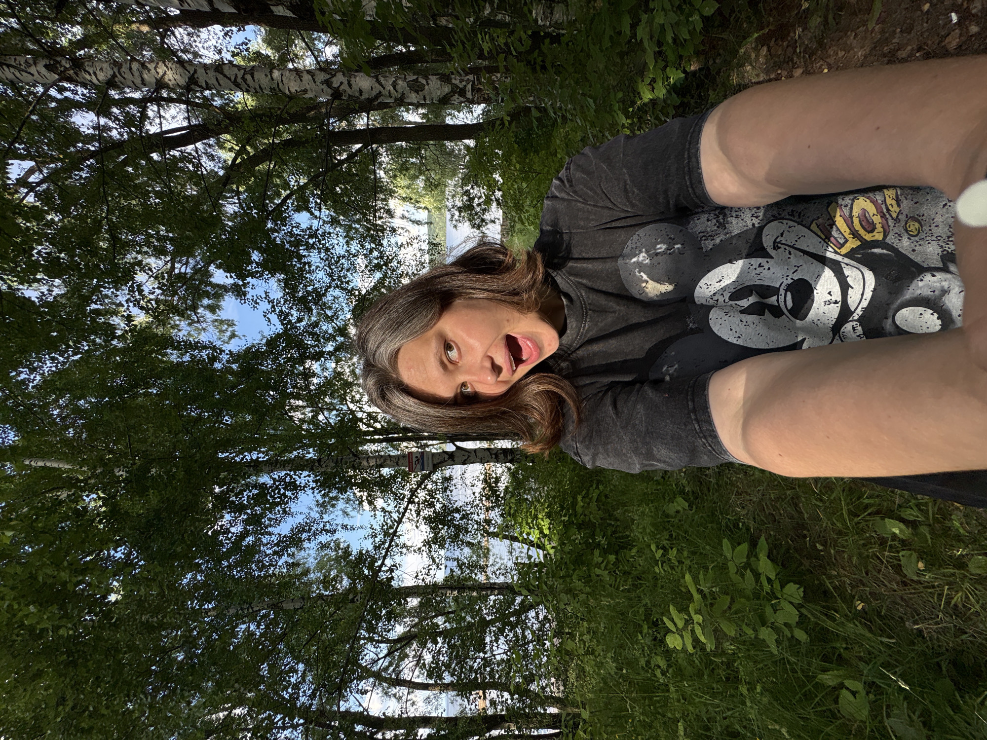 A young woman with medium-length brown hair, streaked with gray, smiles with her mouth open towards the camera. She is wearing a dark grey t-shirt with a faded graphic of Mickey Mouse. Her arms are extended towards the camera, obscuring the lower portion of her body. The woman is outdoors, in a wooded area with tall birch trees. Sunlight filters through the leaves, creating dappled light on the ground. In the background, a body of water, likely a river or lake, is visible through the trees. The overall setting suggests a natural environment, possibly a park or recreational area. The image appears to be a selfie, taken from a low angle. The location is identified as Chaikovsky, Russia.