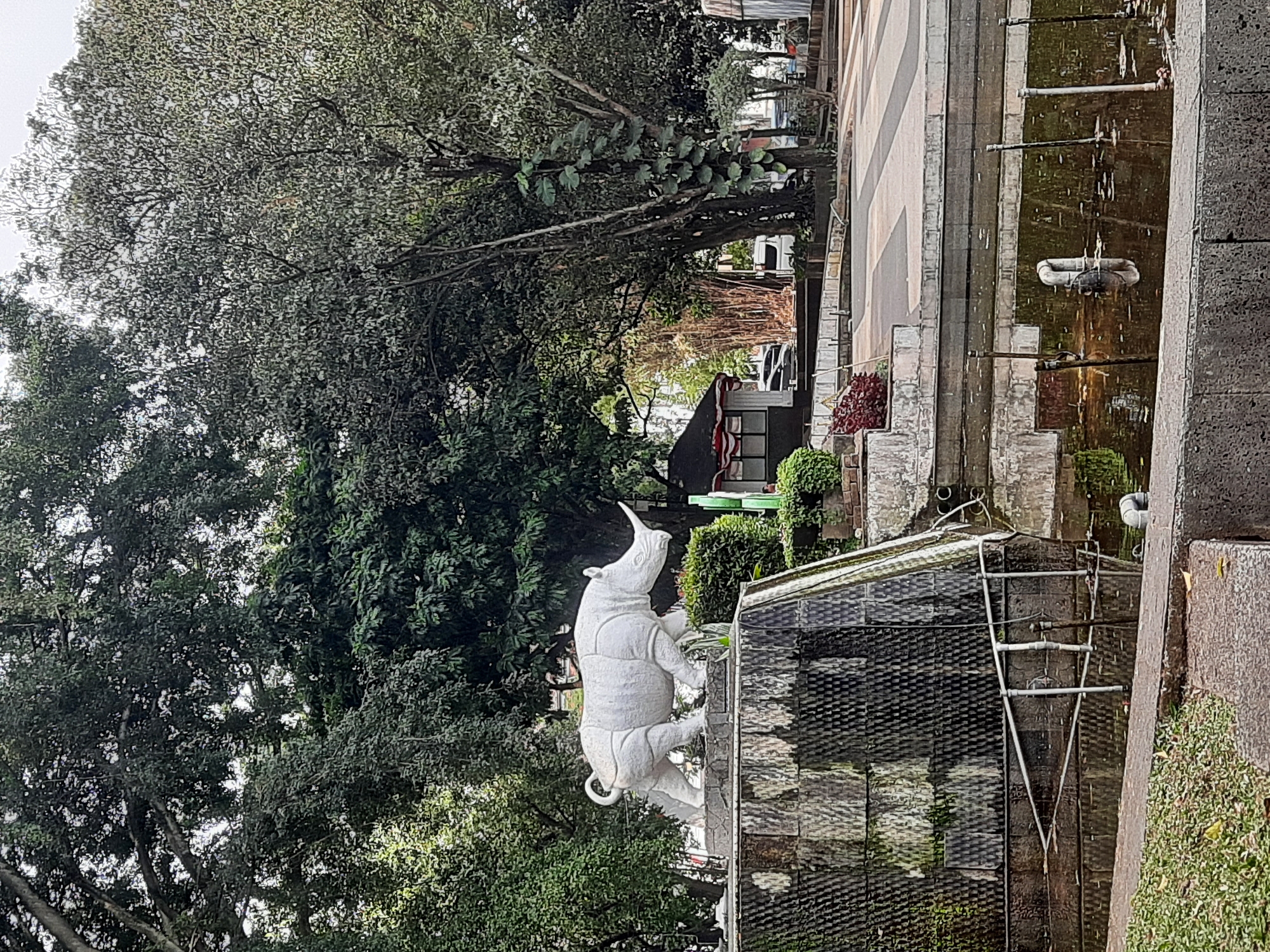 A white stone rhino statue stands on a raised platform with a textured dark gray facade, which is part of a fountain structure. The fountain itself has dark, still water. The background is filled with lush green trees, some with large leaves and vines, and others with denser foliage. In the middle ground, there is a small building with a red and white striped awning. The ground in front of the fountain is paved with gray and light gray geometric patterns. There are no visible people in the image. The scene appears to be an outdoor public space, likely a park or plaza, in Bandung City, Indonesia, given the architectural style of the building and the type of vegetation.