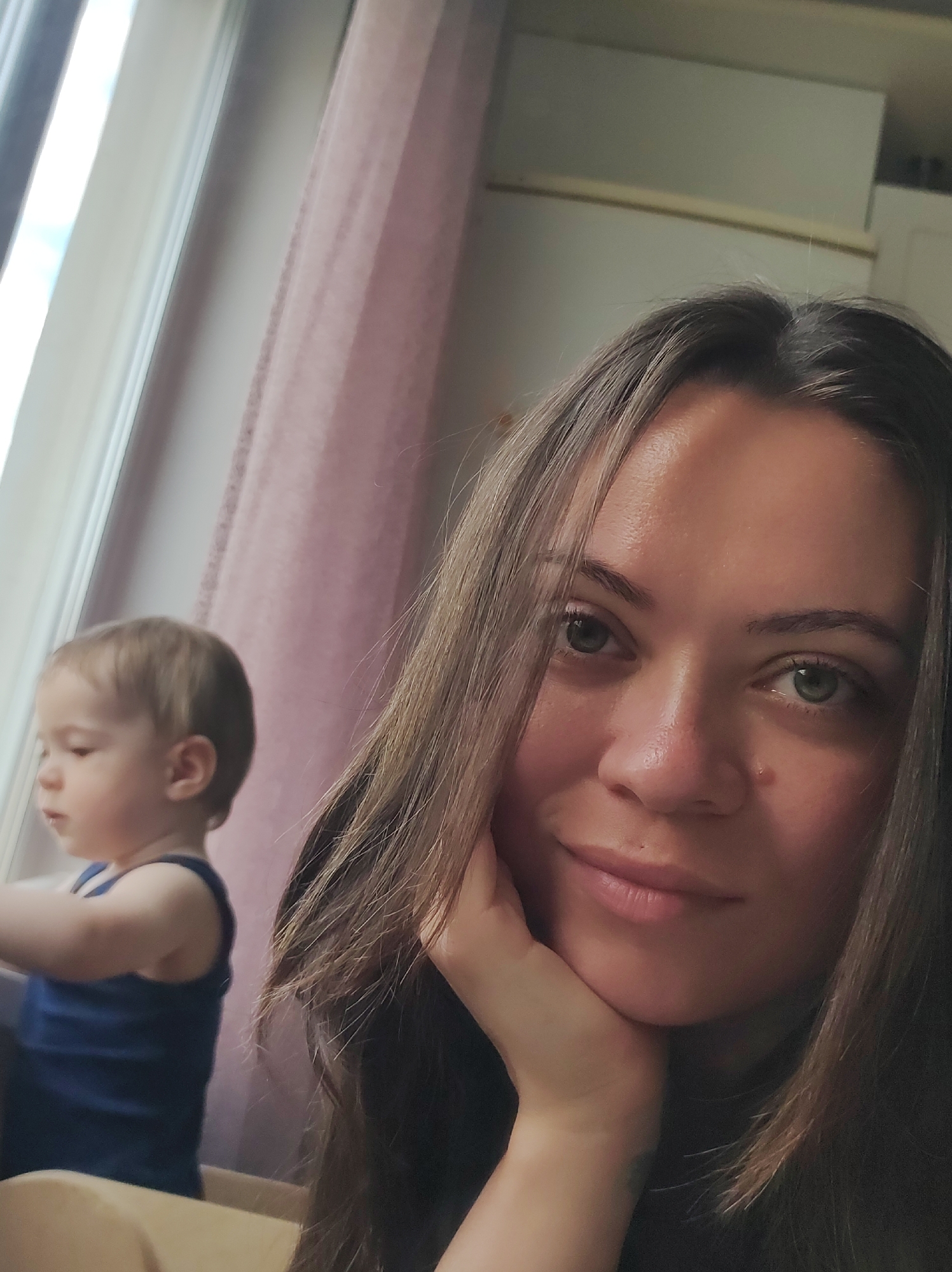 A woman is taking a selfie with a young child near a window. The woman is in the foreground, positioned to the right of the frame. Her head is resting on her hand as she looks toward the camera with a slight smile. She has brown hair and light eyes. To the left, partially visible, is a young child wearing a blue tank top, looking out of the window. A white window frame is prominent to the left, with a translucent pink curtain visible behind it. The background includes what appears to be a cabinet or shelving unit. The location is Chaikovsky, Russia.