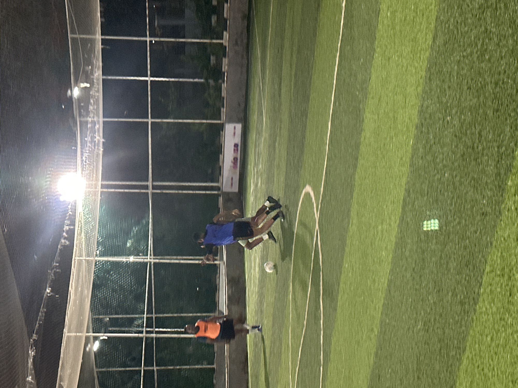 The image depicts an outdoor football pitch with artificial turf, likely a recreational facility. The pitch is illuminated by overhead lights, suggesting the game is taking place at night. Several individuals are visible on the pitch, engaged in playing football. One person, wearing an orange bib and black shorts, stands near the left side of the frame, observing the game. In the center, two individuals are actively playing, with one in a blue jersey and black shorts and another in lighter colored shorts and a dark top. A football is present on the ground between them. The background shows a fence or net enclosure, typical of sports facilities, and trees or foliage beyond. The overall environment suggests a sports complex within a city. While the image itself does not definitively place the location within Abuja, Nigeria, such a facility is consistent with urban recreational infrastructure found in cities like Abuja.