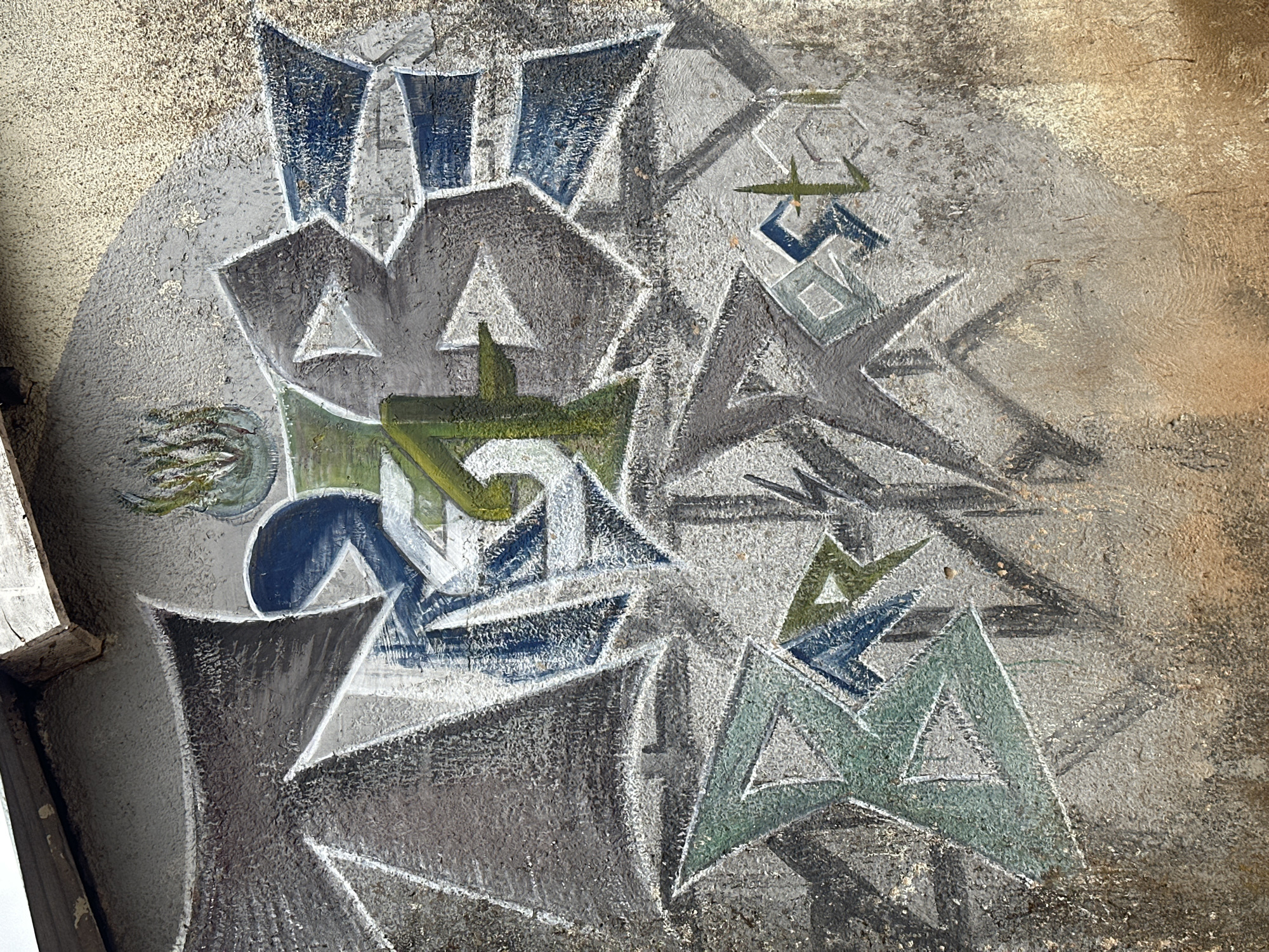An abstract graffiti artwork adorns a textured concrete wall in an urban outdoor environment. The graffiti features sharp, geometric shapes and bold lines in a palette of gray, blue, and green. Central to the composition is a vertical element in green, resembling a stylized plant or abstract structure, with a circular shape above it containing what looks like foliage. Scattered throughout the artwork are angular forms in blue and white, creating a dynamic and somewhat fragmented visual. The wall itself is rough and weathered, with visible imperfections and stains, suggesting an aged urban surface. The lighting indicates a daylight scene, casting subtle shadows and highlighting the texture of the wall and the paint.