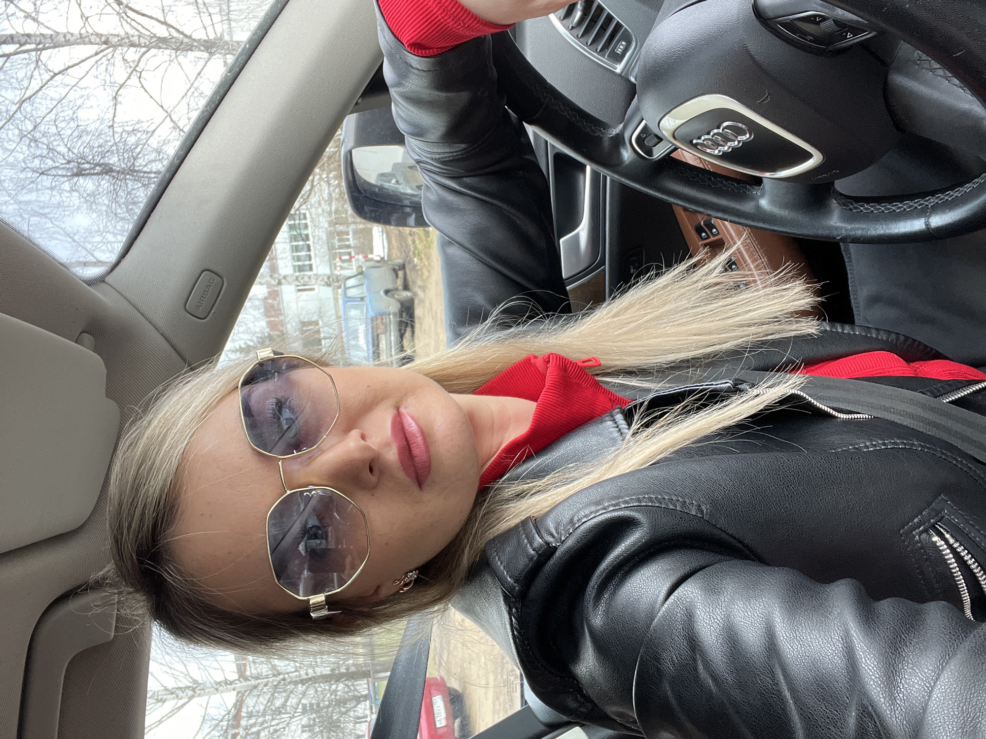 A woman with blond hair and sunglasses is sitting in an Audi car. She is wearing a black leather jacket over a red top. Her left hand is on the steering wheel, which displays the Audi logo. The background shows a street scene, with buildings and vehicles visible through the car windows. The car's interior is gray. Based on the surroundings and the "Airbag" sign in the car, the photograph was most likely taken in the city of Chaikovsky, Russia.