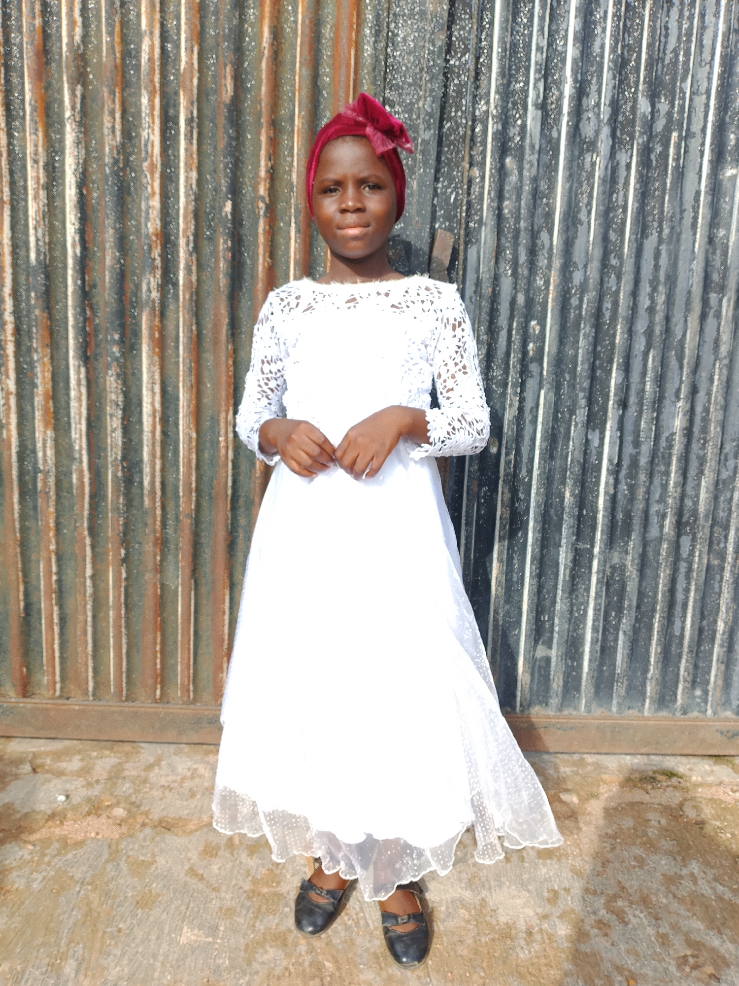 A full-body, slightly low-angle shot shows a young girl standing in front of a corrugated metal fence. She is facing forward, with her hands clasped in front of her waist. The girl is wearing a white lace dress with long sleeves and a white sheer overlay on the skirt. She also has a red headscarf tied around her head. Her feet are clad in black Mary Jane shoes. The background fence is a mix of rust-colored and dark gray corrugated metal panels, with vertical lines running throughout. The ground in front of the fence is a textured concrete surface, with some dirt and debris visible. The lighting suggests it is daytime, with sunlight casting shadows.