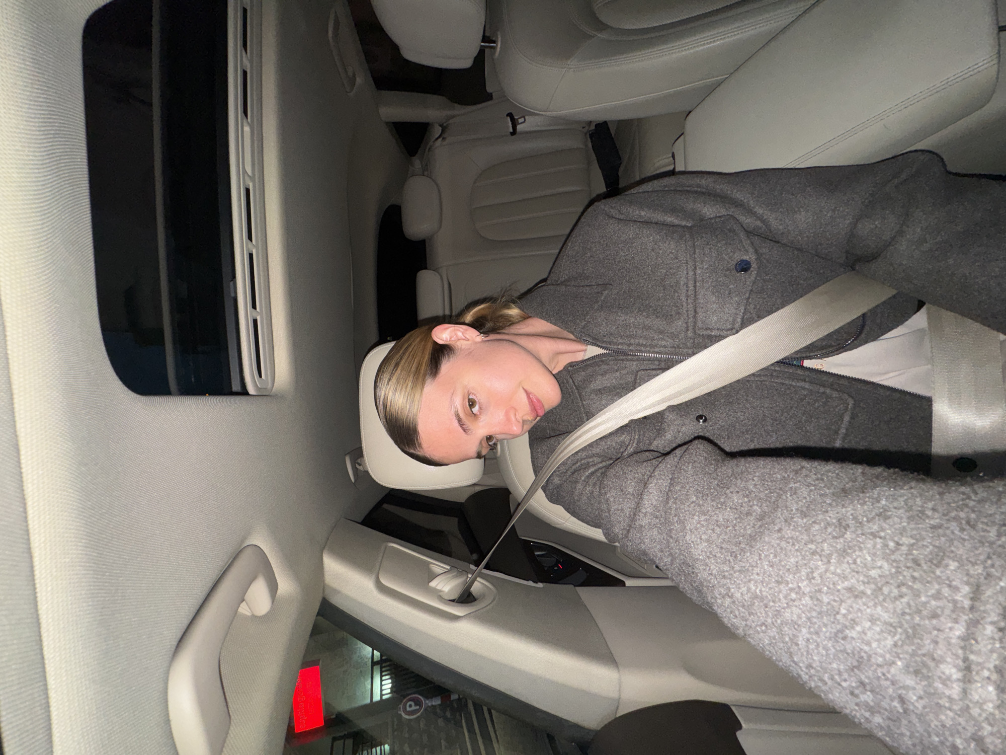 A woman with light skin and blonde hair is sitting in the passenger seat of a car. She is wearing a gray coat and a white shirt and has a seatbelt fastened across her body. The car has gray leather seats and a sunroof. Outside the car, there is a red sign and a parking sign. The location is a parking garage or street parking area. The city is not possible to infer from the image.