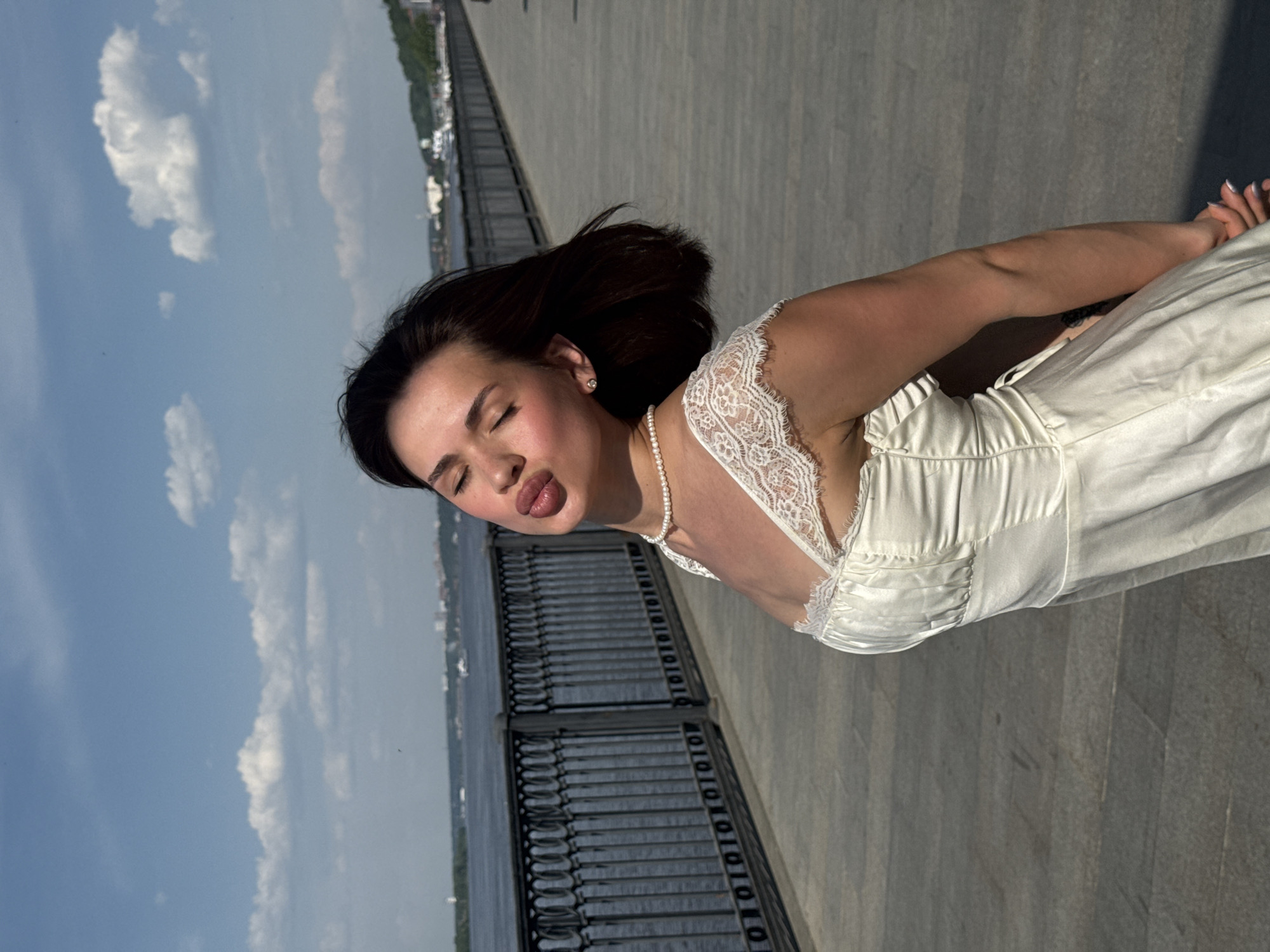 The image depicts a woman in a white dress standing on a paved walkway in Perm, Russia, next to a metal fence overlooking a river. She has dark hair and is wearing a pearl necklace. The sky is partly cloudy.