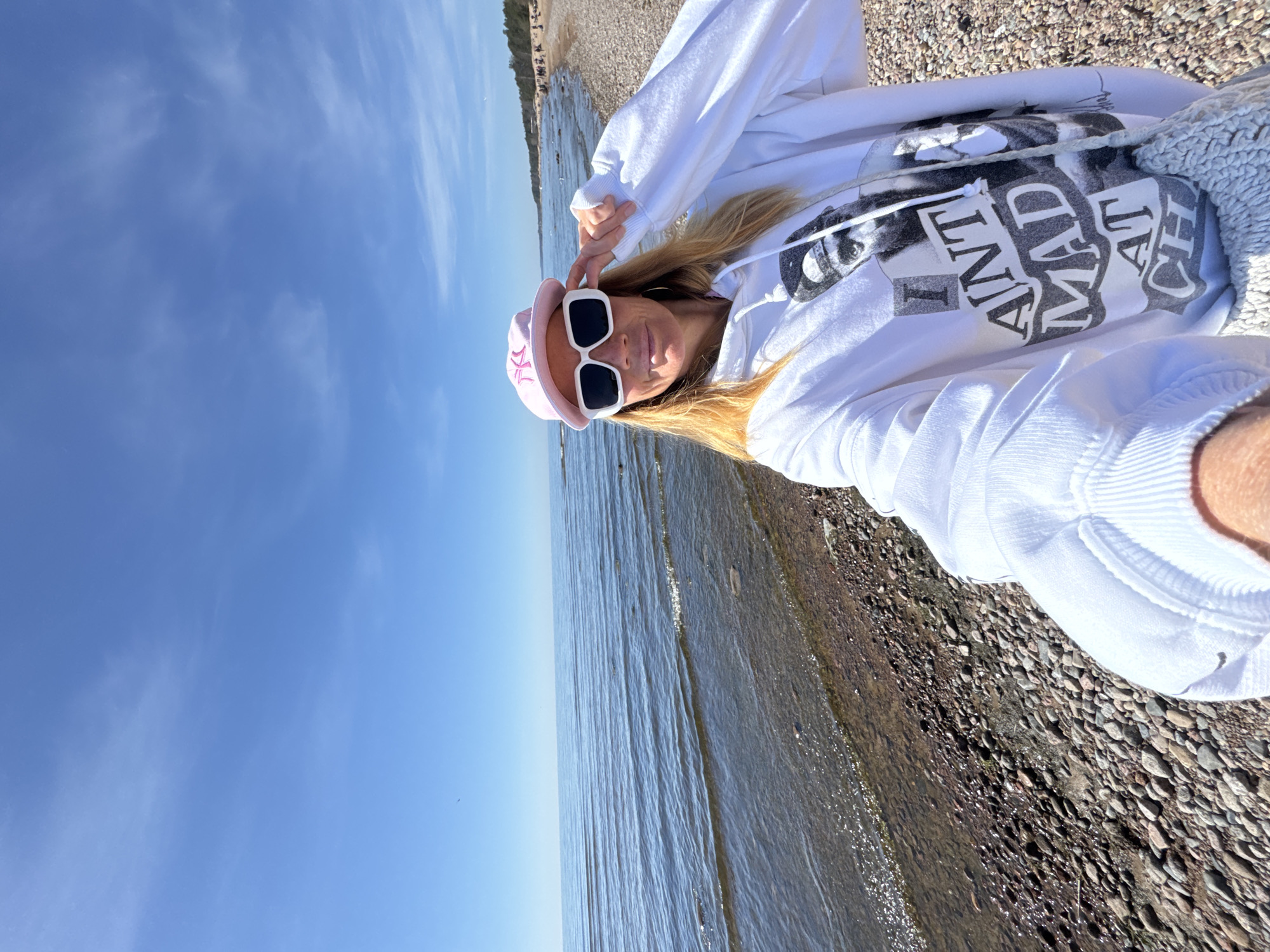 A woman is taking a selfie at the beach in Komarovo, Russia, wearing a pink hat, white sunglasses, and a white hoodie with a graphic print. She is standing on a pebble beach, with the sea on one side and a clear blue sky above. The camera is angled downwards, capturing her face and upper body.