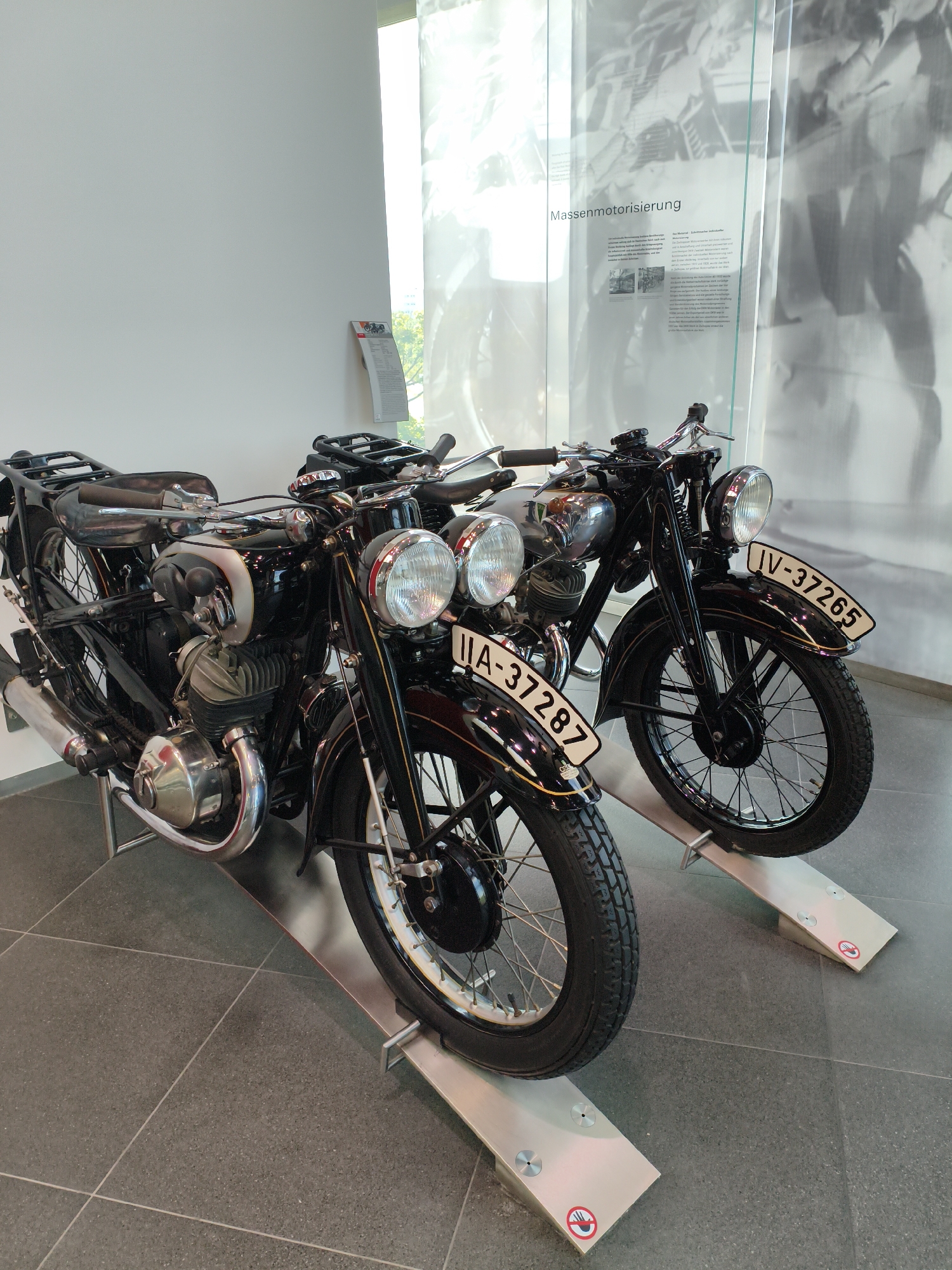 Two vintage black motorcycles are on display indoors. The motorcycle in the foreground has a license plate that reads "IIA-37287". The motorcycle in the background has a license plate that reads "IV-37265". Both motorcycles are positioned on metal ramps. Behind the motorcycles, there is a large glass panel with text and images displayed on it. The text on the glass panel appears to be in German. The environment appears to be a museum or exhibition space, with a light-colored wall in the background and a tiled floor. The lighting suggests an indoor setting, possibly with natural light coming from a window or skylight. The location is identified as the Audi Forum in Ingolstadt, Germany.