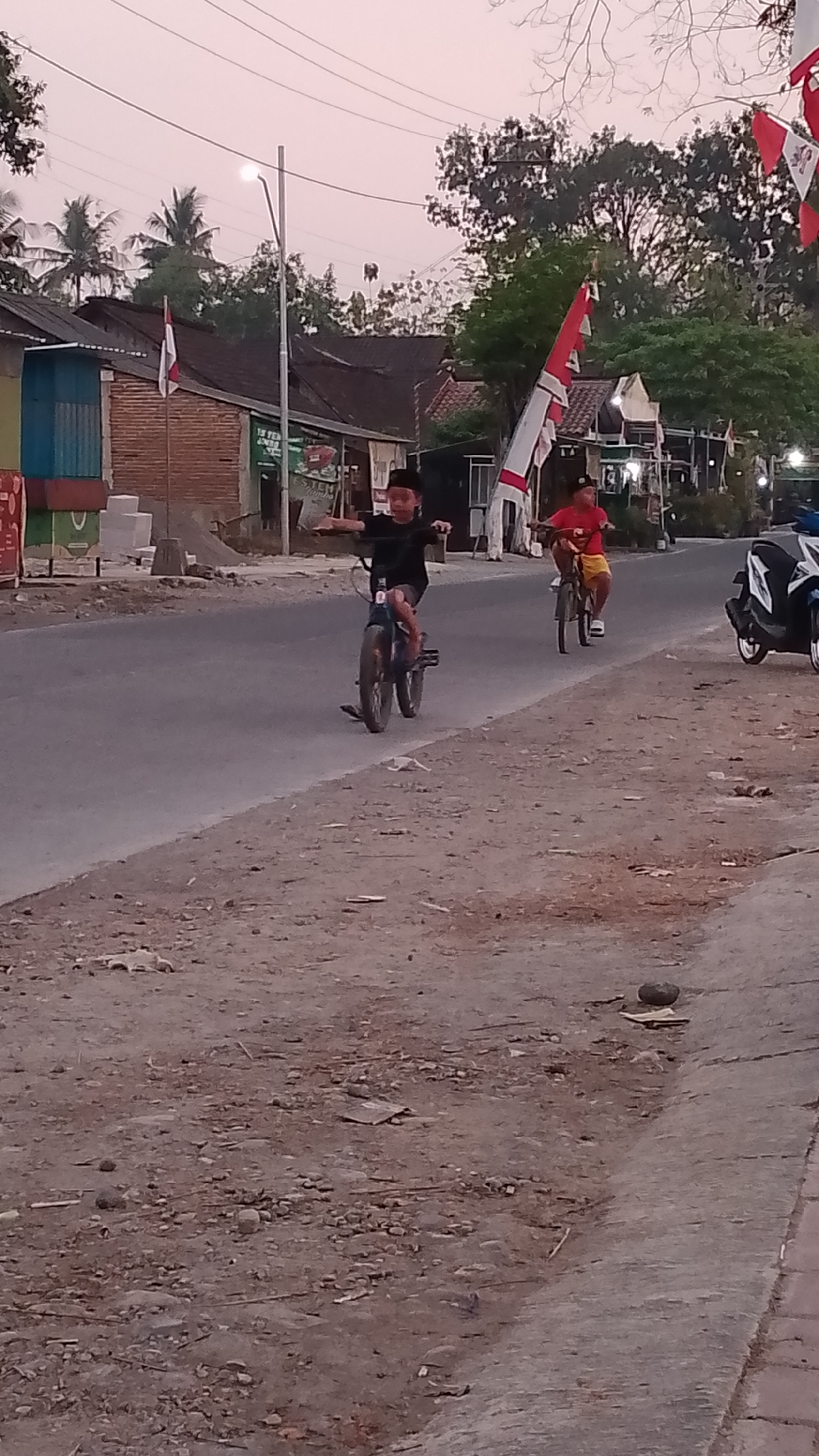 The image shows two young boys riding bicycles on a paved road. The boy on the left is wearing a black shirt and black pants and is riding a dark-colored bicycle. The boy on the right is wearing a red shirt and yellow shorts and is riding a bicycle. There are buildings and trees in the background. Indonesian flags are visible on some of the buildings, suggesting the location is Karanglangu, Indonesia. A scooter is parked on the right side of the road. The sky is a pale pink, indicating it is either dawn or dusk. The ground on the side of the road is unpaved and covered with dirt, rocks, and debris.