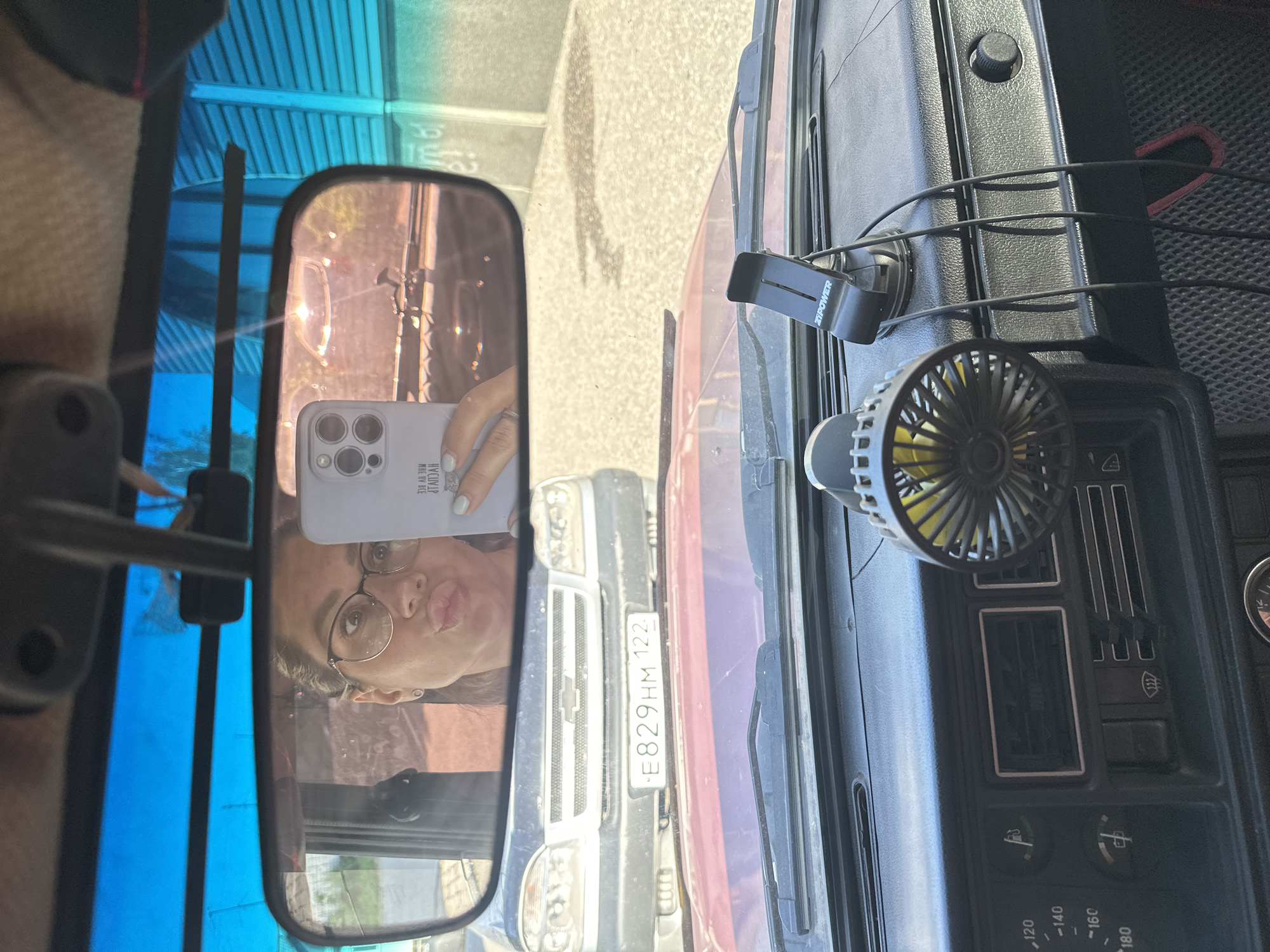 The image shows the interior of a car, with a rear-view mirror reflecting a woman taking a selfie. She is wearing glasses and making a kissing face, holding a smartphone. Outside the car, another car with a license plate reading "E829HM 122" is visible. The dashboard of the car features a small fan and a phone holder. The context suggests this is likely a residential street in Novoaltaysk, Russia, given the car's license plate format.