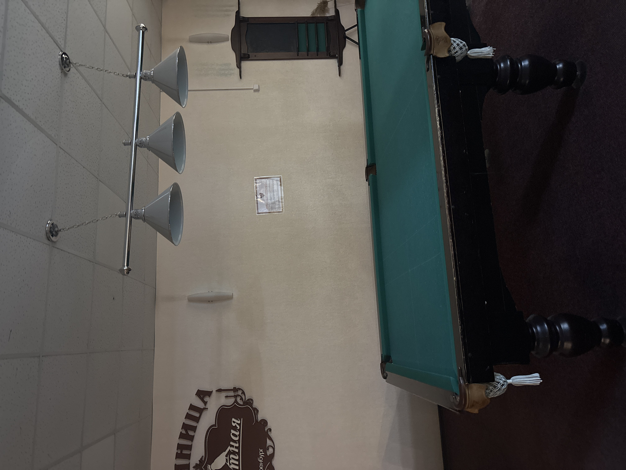 The image depicts a pool hall, likely in Orenburg, Russia, based on the Cyrillic text on the wall. A green-felted pool table with dark wood trim and decorative tassel details is positioned in the center of the frame, partially obscured by darkness. A light fixture with three downward-facing cone-shaped shades hangs from the ceiling, illuminating the table. A dark wooden cue rack is visible on the wall above the table. The wall is off-white and has a small framed certificate.