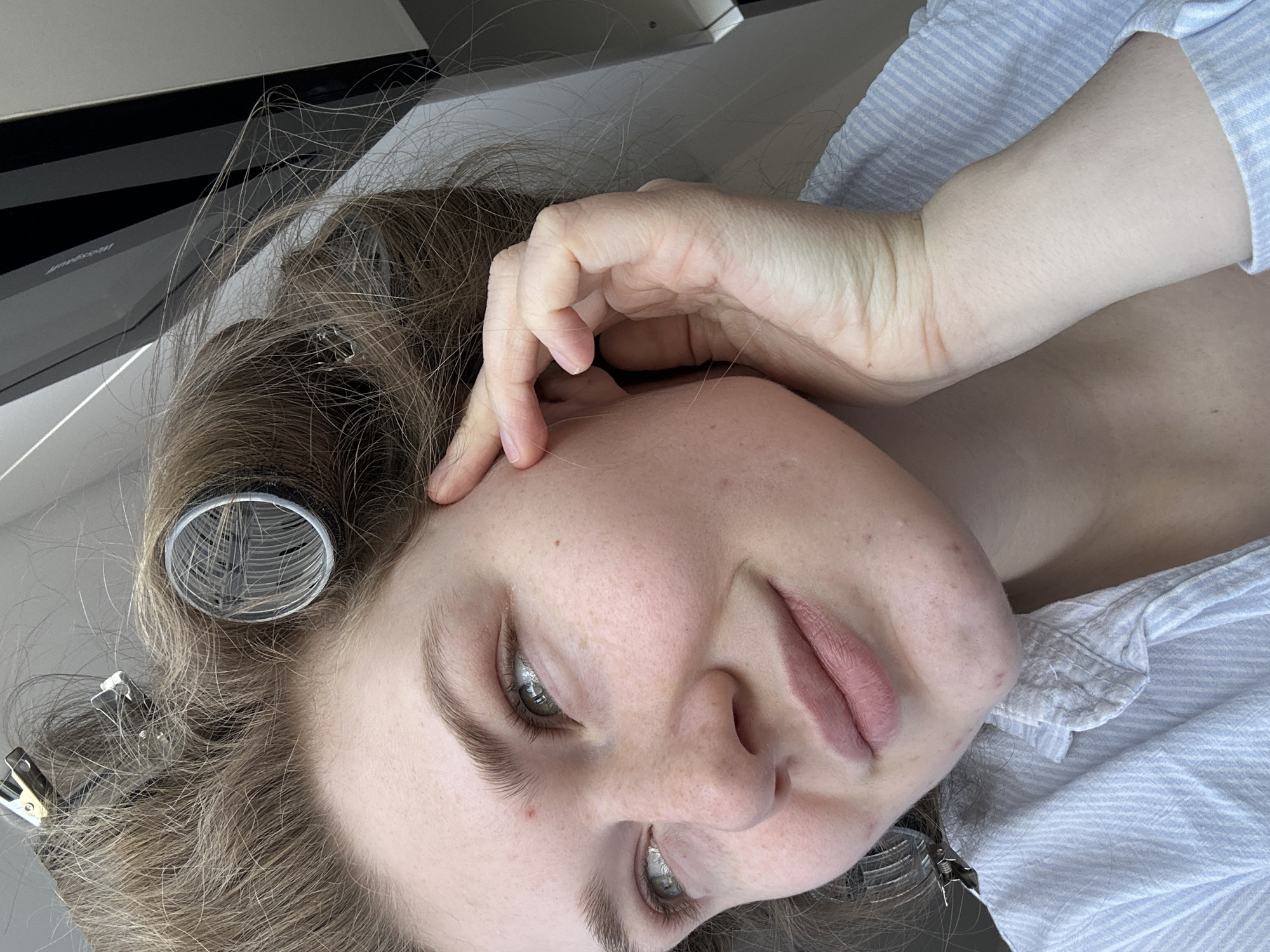 A close-up, angled shot depicts a woman with light skin, likely indoors. Her hair is brown and styled with curlers and metal clips. She rests her fingers on her temple, and her facial expression suggests relaxation or contemplation. She is wearing a light blue striped shirt. The background includes a white surface and what appears to be a black appliance, possibly a microwave or oven. There is no indication in the image that the location is Kazan, Russia.