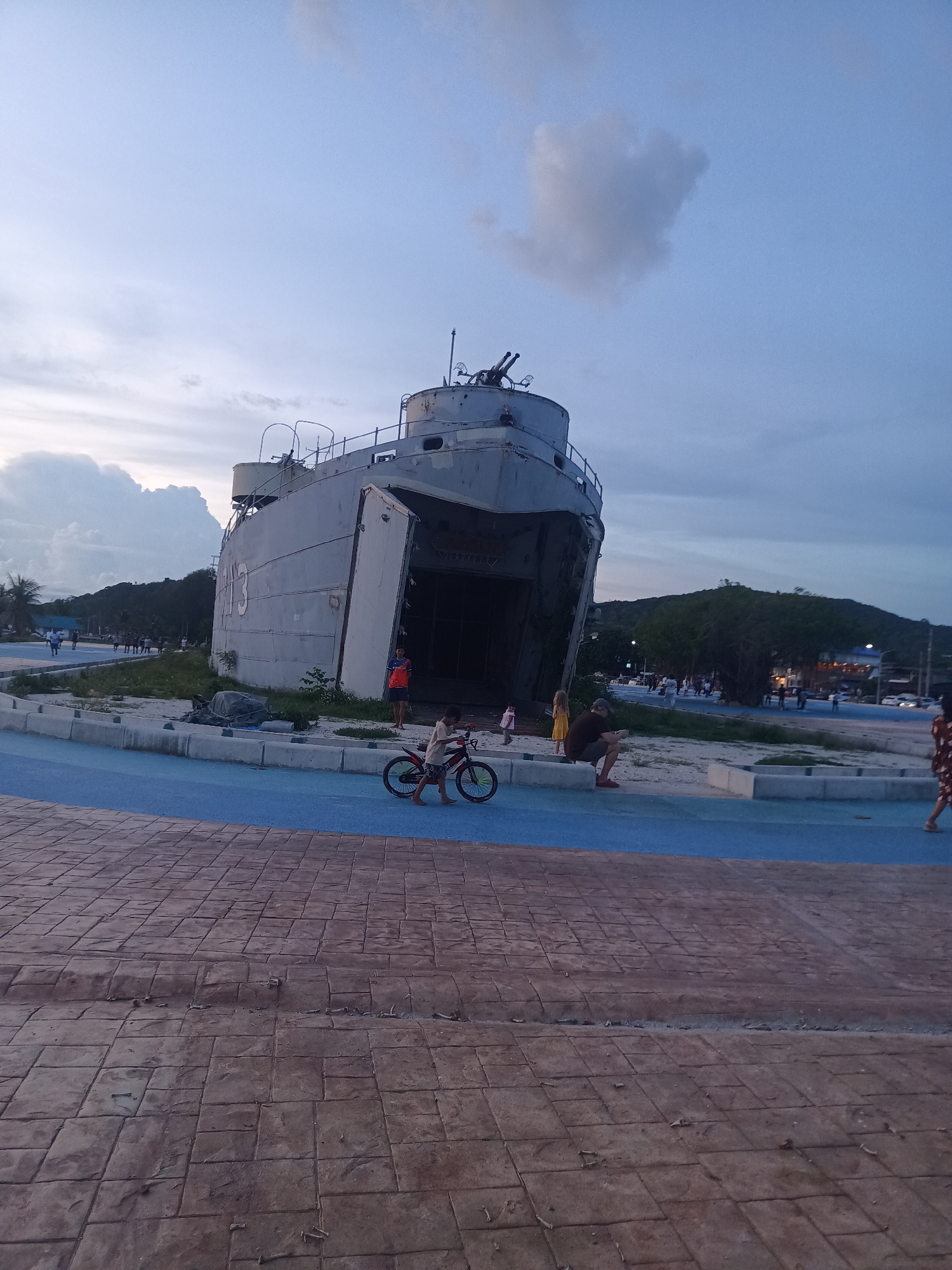 In the city of ตำบลเกาะพะงัน, Thailand, a decommissioned military ship rests on land, serving as a landmark. The sky above is a gradient of twilight blue and gray, with a solitary cloud in the shape of a heart. A boy rides a bicycle on a blue-colored walkway in the foreground, his bare feet pushing pedals. To the right, a man sits on a concrete barrier, looking at his phone. In the background, several people are visible near the ship and walking along a path lined with palm trees. A lush green hillside rises behind the structures, dotted with buildings and lights. The ground is paved with textured, reddish-brown rectangular tiles.