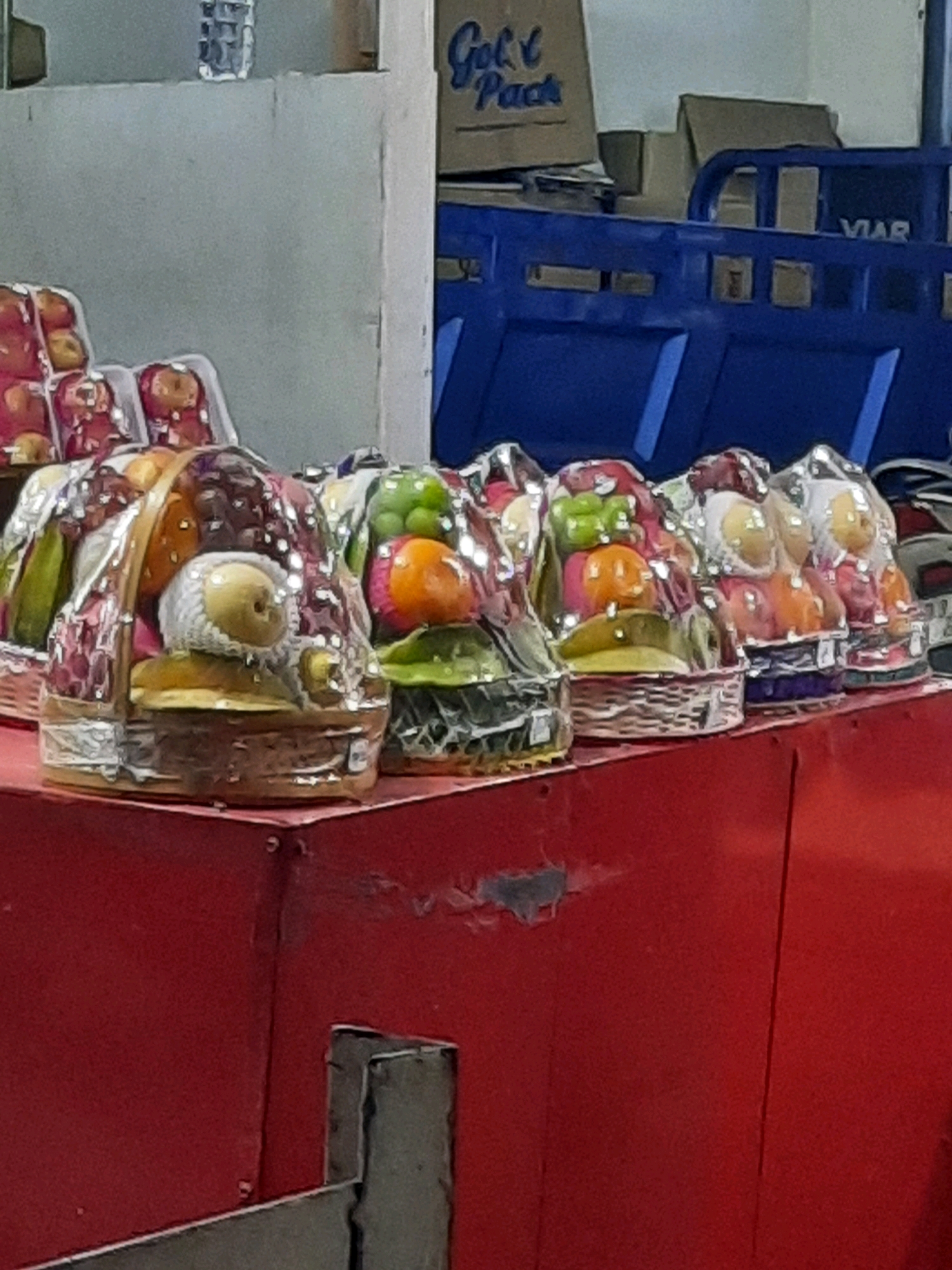 The image captures a close-up, low-angle view of several fruit baskets wrapped in clear plastic, displayed on a red surface. The baskets appear to be of various sizes and contain an assortment of fruits, including apples, pears, grapes, and possibly oranges and bell peppers. The fruits are neatly arranged within wicker or woven baskets, and the plastic wrapping gives them a shiny, preserved appearance.
In the background, there's a light blue rack or shelving unit, with some cardboard boxes stacked on top of it. One box has "Gold Pack" written on it in blue lettering. The overall environment suggests a market stall or a retail display, likely in East Jakarta, Indonesia, given the context. The lighting appears to be artificial, creating highlights on the plastic wrap and the fruits. The red surface in the foreground shows some signs of wear and tear, with scuffs and minor damage. There are no people visible in the image.