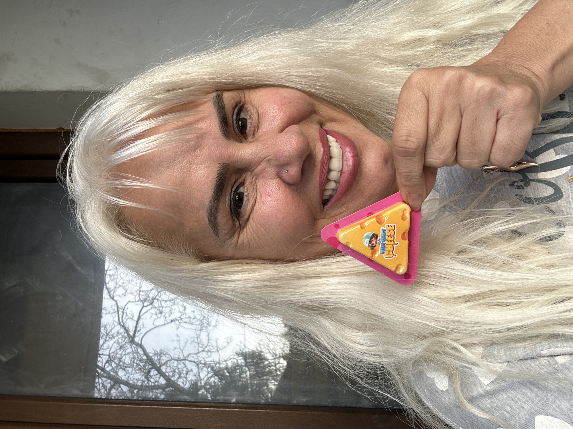 A woman with long, platinum blonde hair is holding a triangular piece of "Ricky Zoom" branded cheese. She is smiling and has a nose piercing. The background shows a window with a tree outside. The location cannot be determined as the photograph does not contain that information.