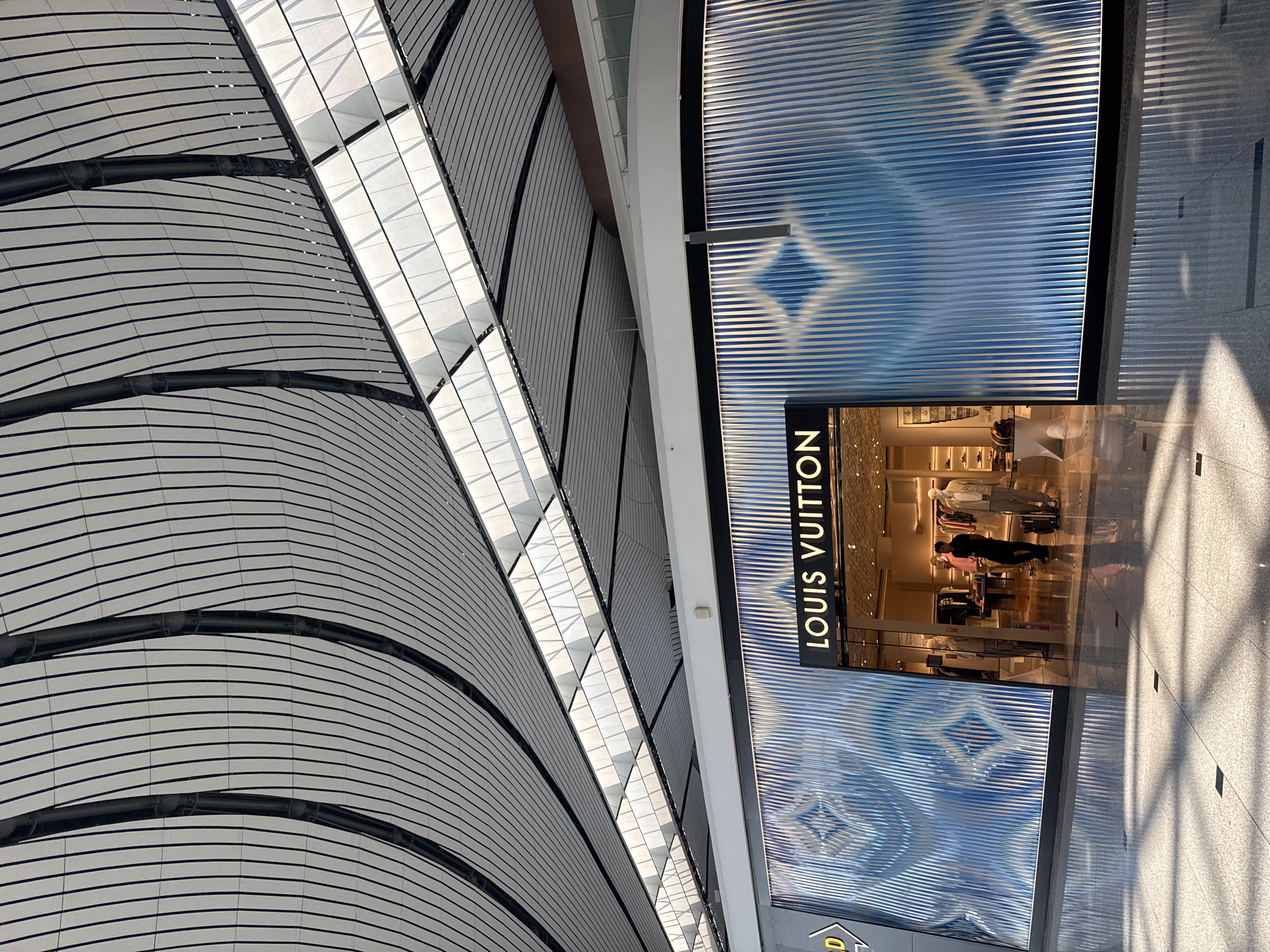 The image shows the interior of a modern airport terminal in Guangyang District, China, with a Louis Vuitton store as the central focus. The store features a striking facade with a blue, diamond-patterned design set against vertical ribbed elements. Above the store, the ceiling is a complex architectural design of white curved panels separated by dark, curved beams, with rectangular skylights allowing natural light to filter in.
Inside the store, several mannequins are dressed in light-colored clothing, and display shelves are visible. A male store employee, wearing a black shirt and dark trousers, stands near the entrance, holding a bag and looking towards the interior. The polished floor reflects the lighting and the architectural elements of the ceiling and store. The overall scene suggests a high-end retail environment within a contemporary airport setting.