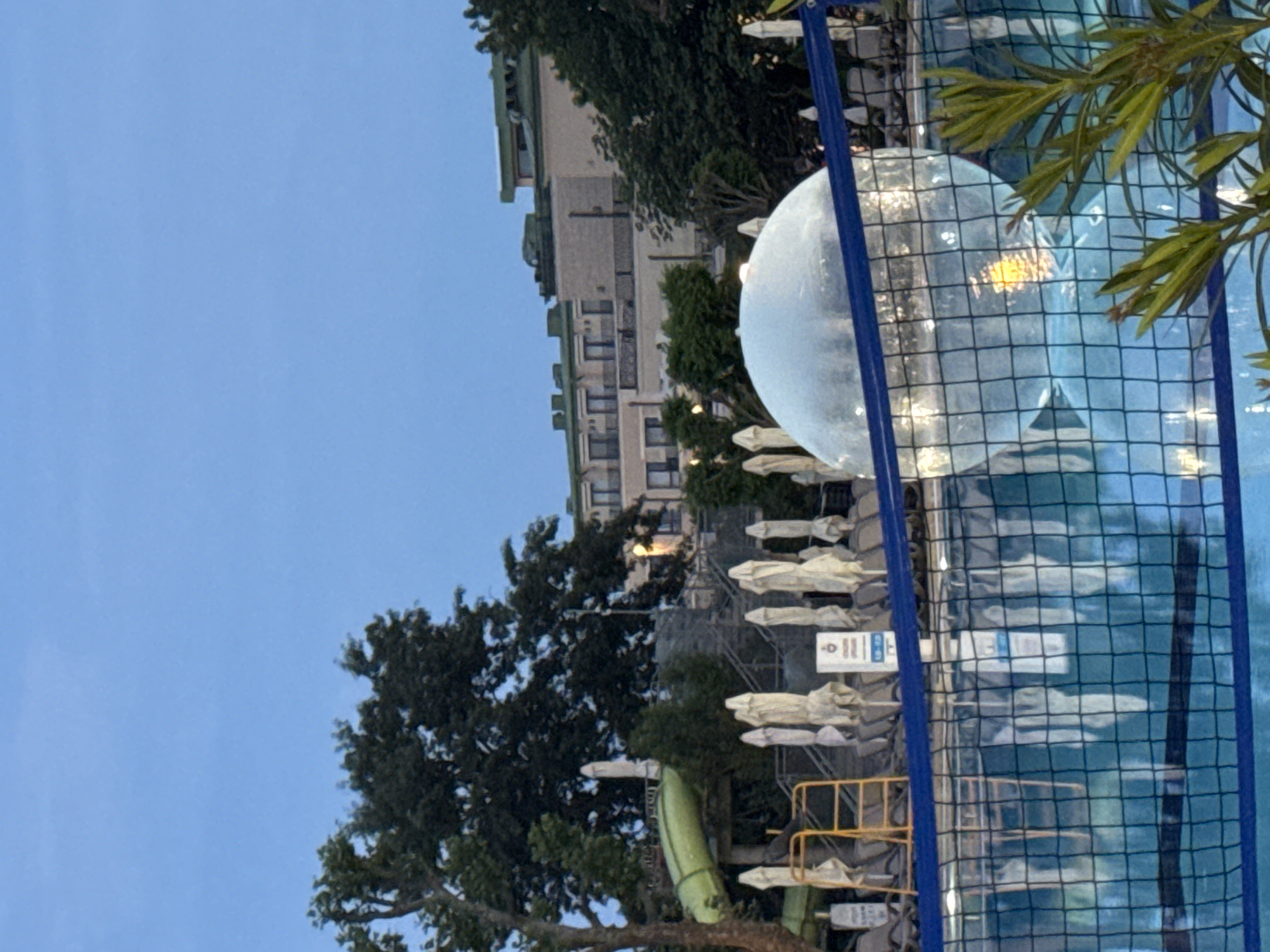 The image depicts an outdoor swimming pool area, likely belonging to a hotel or resort, under a clear, light blue sky. In the background, a multi-story building with numerous windows and what appear to be balconies is partially visible through green trees and foliage.
The main subjects in the foreground are a large, translucent spherical object floating in the blue water of the swimming pool, and a blue net that extends across the frame, partially obscuring the pool and the background. Alongside the pool, a row of white folded sun loungers or umbrellas is neatly arranged. To the left, a green inflatable slide or structure is present, nestled among more trees. A yellow metal railing or barrier is also visible near the sun loungers.
The scene suggests a leisurely setting, though no specific activity like swimming or sunbathing is clearly depicted, and no people are visible. The lighting, with its soft tones and clear sky, indicates it is daytime, possibly late afternoon or early evening. There are a few white signs with dark text near the sun loungers, but the text is too blurry to be legible. The overall impression is one of tranquility in an outdoor recreational space.