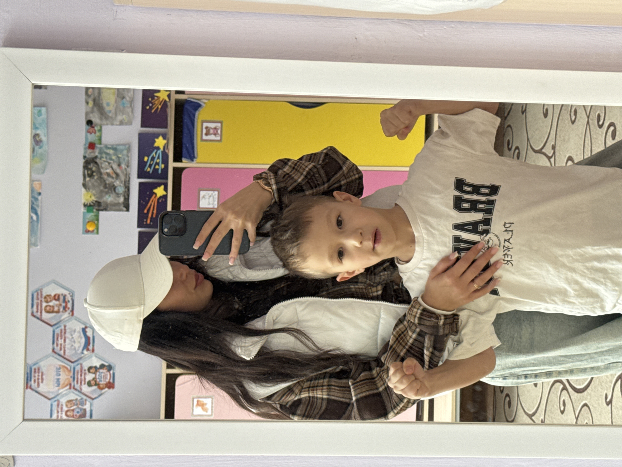 A reflection in a large mirror shows a woman in a white baseball cap, white vest, and brown plaid shirt holding up her cell phone while embracing a young boy wearing a white t-shirt with large black letters. The reflection also reveals a set of shelves with brightly colored drawers behind them. Artwork and decorations are visible on the wall to the left of the shelves. The image appears to be taken inside a room in a location in the city of Chaikovsky, Russia.