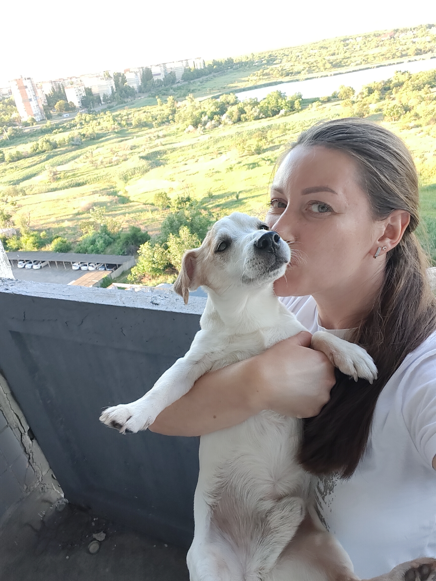 A selfie is taken by a woman holding a white and tan dog on a balcony overlooking a cityscape and a natural landscape. The woman is positioned in the right half of the frame, her face turned towards the dog, leaning in to kiss its cheek. She has long brown hair and is wearing a white t-shirt. Her left arm is wrapped around the dog, supporting its body, while her right hand is holding its hind legs. The dog, facing left, has its head tilted upwards towards the woman's face, with its eyes open.
The background consists of a panoramic view of Kryvyi Rih, Ukraine. In the middle ground, there are several multi-story apartment buildings and residential structures. Below and to the left of the buildings, a covered parking area with several cars is visible. Beyond the cityscape, a vast green landscape unfolds, with rolling hills and vegetation, and a body of water, possibly a river or reservoir, can be seen in the distance. The sky above is bright, indicating daylight. The balcony railing is in the foreground, and parts of the building's exterior, with visible cracks and discoloration, are also present.