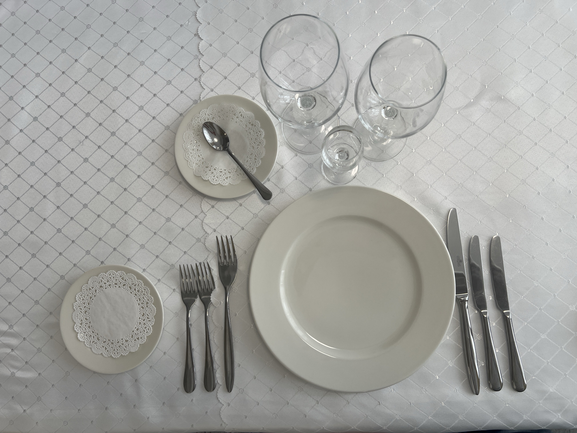 The image displays a meticulously set dining table for one person, viewed from an overhead perspective. A pristine white tablecloth, featuring a subtle, repeating diamond-shaped pattern, covers the surface.
At the center of the setting is a large, round, cream-colored dinner plate. To the left of this main plate, three silver forks of differing sizes are arranged, with the largest closest to the plate. To the right, three silver knives are neatly aligned, with the largest knife also closest to the plate. One of the knives clearly shows the brand "DUSLAR" etched into its blade, while another appears to have similar, though less distinct, text.
In the upper-left quadrant of the frame, a smaller side plate holds a single silver spoon, which rests upon a white, circular paper doily with a delicate, lace-like edge. Another identical small plate with a matching white paper doily is positioned in the lower-left corner, likely intended for bread or an appetizer.
Towards the upper-right area of the table, three clear glass goblets of varying heights and capacities are arranged. Two appear to be wine or water glasses, while the smallest is a shot or cordial glass.
The overall scene depicts a formal place setting, indicative of a prepared meal with multiple courses, likely in a restaurant, hotel, or event setting in Votkinsk, Russia. No people are visible, and no specific time of day or weather conditions can be discerned from the image.