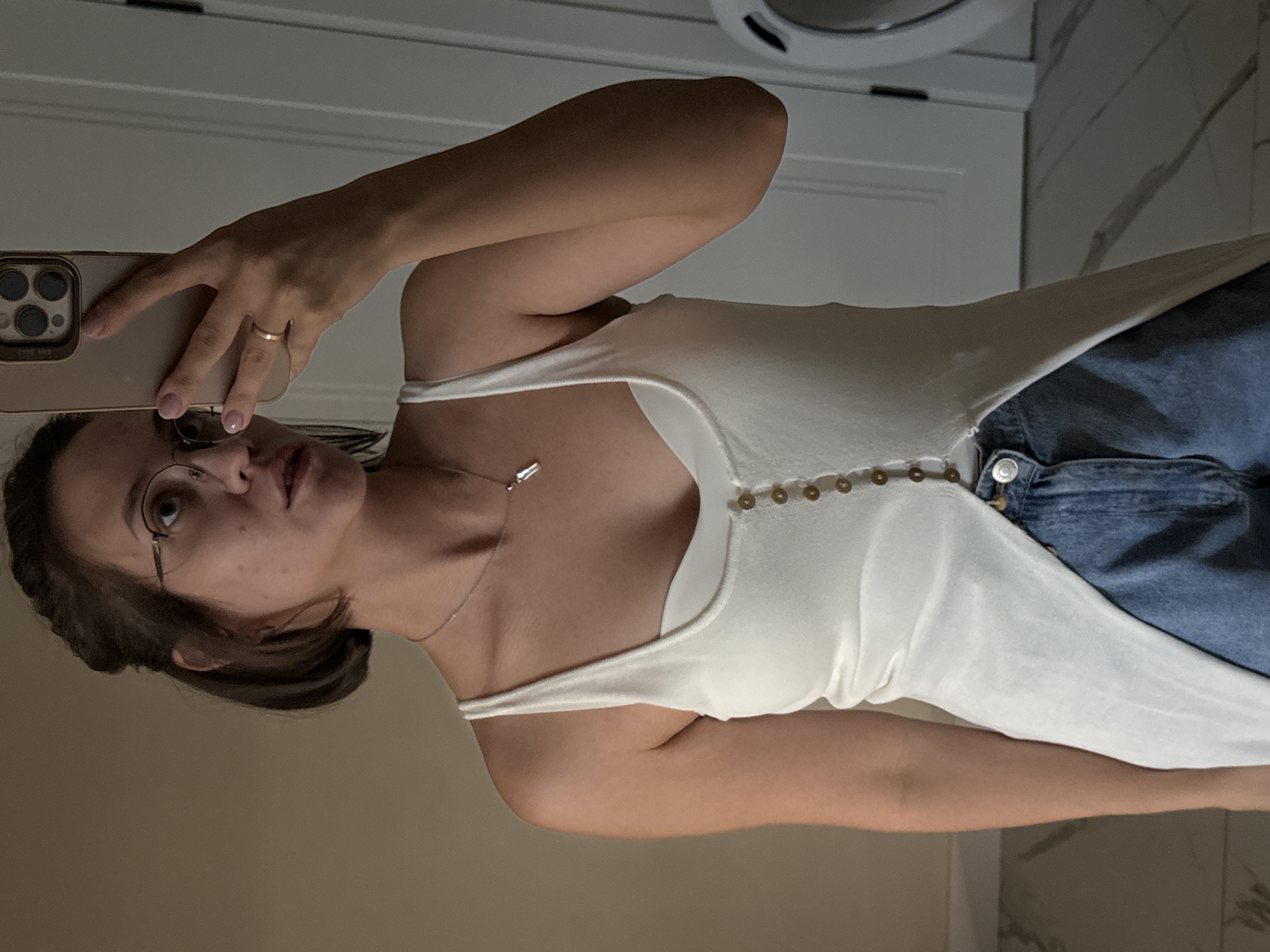 A woman with short, dark hair pulled back, wearing round glasses and a white tank top over a white camisole, is taking a selfie in what appears to be a bathroom. She is holding a gold-colored smartphone in her right hand, capturing her reflection in a mirror. Her left arm is visible, resting at her side. She is wearing a delicate silver necklace with a clear pendant and a thin gold band on her left ring finger. Her lower body is clad in blue denim jeans. The background shows a closed white door and a white washing machine on the right side. The floor has a tiled pattern.