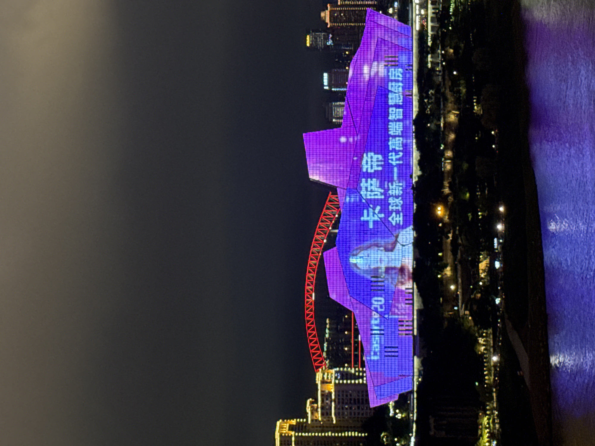 This is a nighttime photo of a modern building illuminated by vibrant purple lights and digital projections. The building has a unique, angular architectural design, resembling a faceted gemstone. Projected onto its surface are Chinese characters and what appears to be a stylized image of a person. Below the main illuminated structure, other buildings with visible yellow lights suggest an urban setting at night. A red Ferris wheel-like structure is also partially visible, adding another element of entertainment or landmark to the scene. The surrounding area is dark, with trees and distant lights indicating a park or waterfront area. The reflection of the purple lights on a dark, rippling surface, likely water, suggests the building is situated near a river or body of water. The text visible on the building reads "卡萨帝 一代高端智慧厨房" and "CASARTE 2011.02.23". The overall impression is one of a dynamic and visually striking urban nightscape in Yuzhong District, China, likely part of an event or celebration given the elaborate lighting and projections.