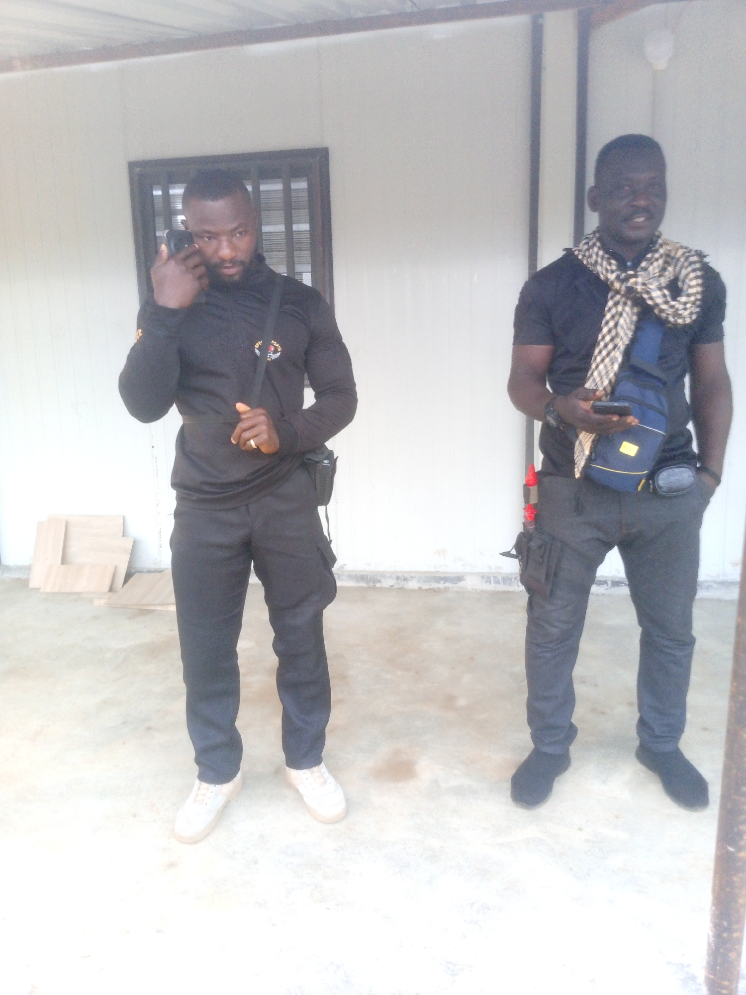 Two men are standing in front of a white, corrugated metal wall. The man on the left is wearing a black long-sleeved shirt and black pants. He is holding a black cell phone to his ear with his left hand and has a small black bag slung across his body. He is also wearing white sneakers. The man on the right is wearing a black t-shirt and black pants. He has a checkered scarf around his neck and a blue and yellow sling bag across his chest. He is holding a black cell phone in his right hand. The ground is concrete and there are some wooden planks stacked to the left of the men. The scene appears to be outdoors. The location is identified as Indo, Nigeria.