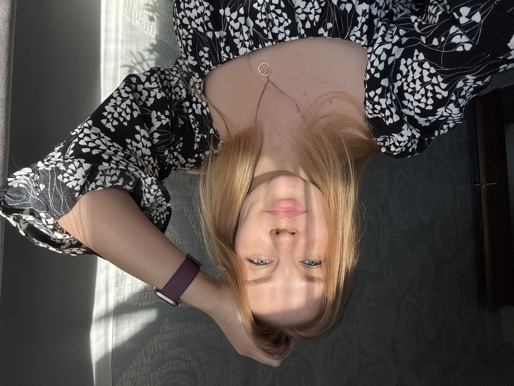A young woman with fair skin and light brown hair is facing the camera. Her head is tilted to the side and she is holding it with her left hand, on which she is wearing a purple smartwatch. She is wearing a black and white floral print dress and a gold necklace with a circular pendant. The lighting is bright, casting shadows across her face and the background. The background appears to be a wall with a subtle pattern and a window to the left. The specific location in the city of Rusanovo, Russia is not discernible from the image.