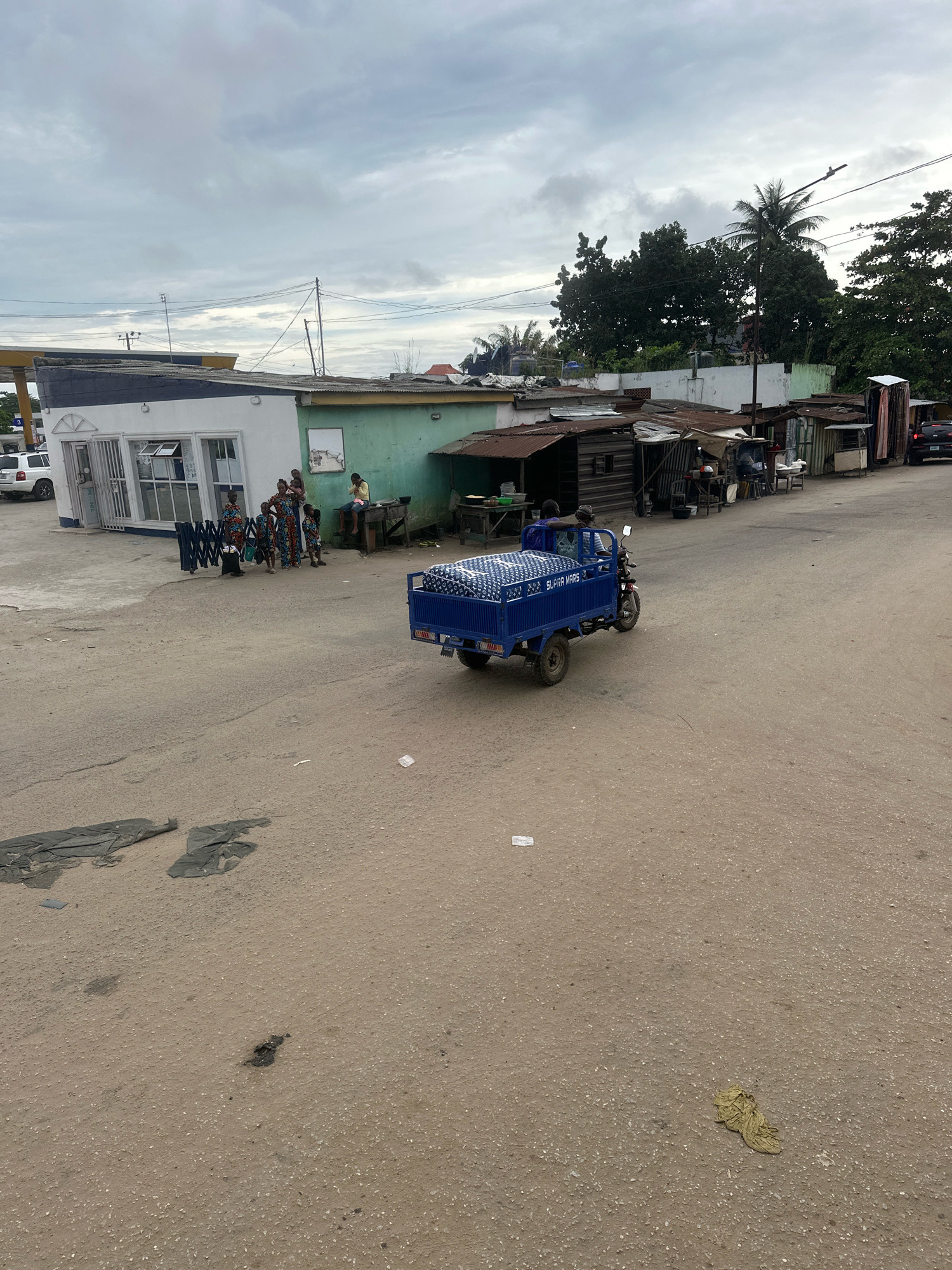 A blue three-wheeled motorized cargo tricycle, referred to as a "motortuk" or "okada," is driving on a paved road in Ajara, Nigeria. The tricycle's cargo bed is filled with stacked mattresses. A man is operating the tricycle, and another person is riding as a passenger. In the background, a white building with large windows and blue trim is visible, potentially a shop or business. Adjacent to it are several smaller, tin-roofed structures that appear to be informal market stalls or dwellings. A group of people, including women and children wearing vibrantly colored traditional clothing, are standing near the white building. The overall environment suggests a bustling, semi-urban setting with a mix of commercial and residential structures. The sky is overcast with clouds.