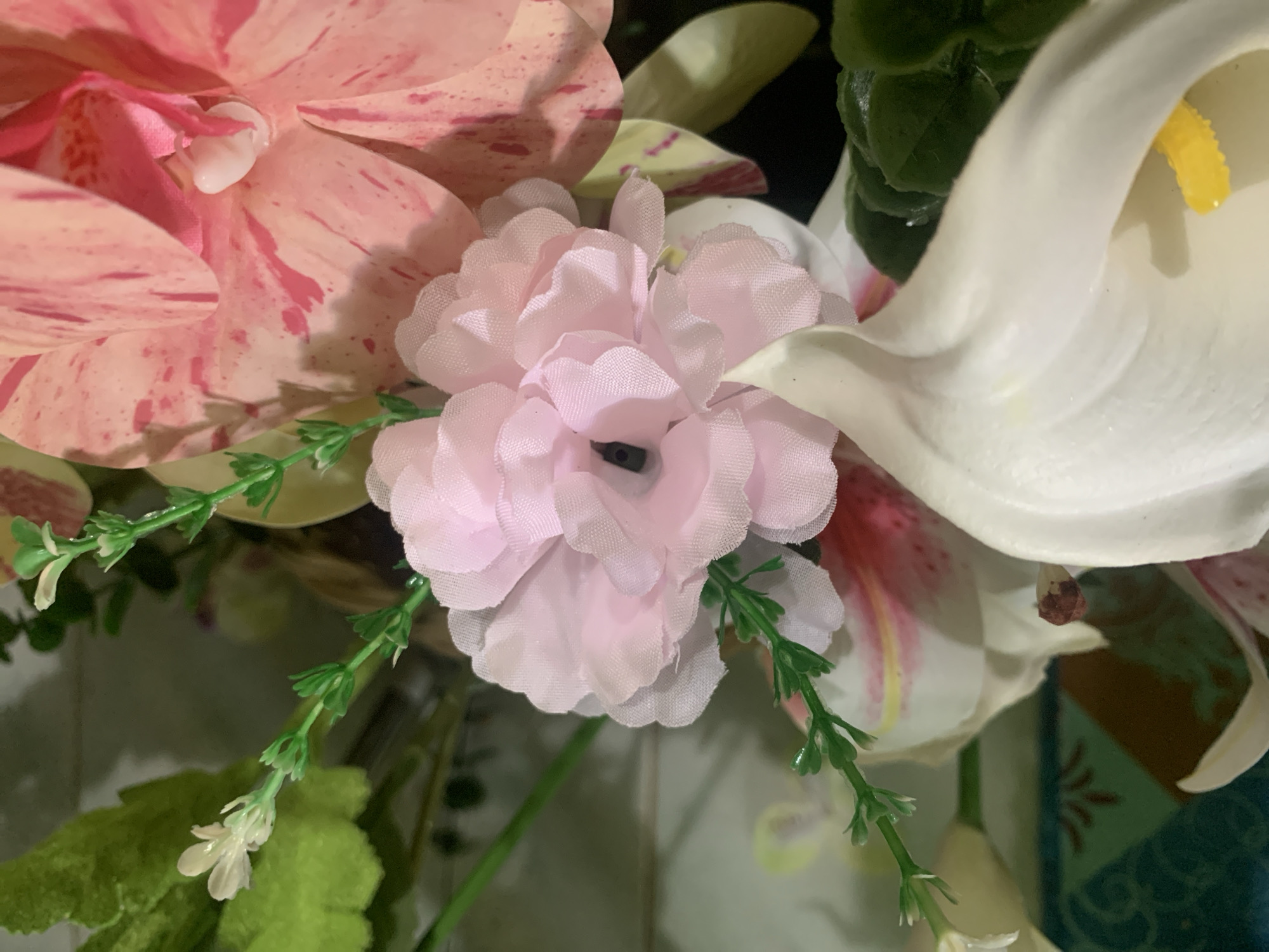 A close-up, eye-level shot showcases a vibrant arrangement of artificial flowers. In the foreground, a delicate pink flower with multiple ruffled petals is centrally placed, its dark center hinting at its artificial nature. To the right, a large, pink orchid with striking red speckles commands attention, its petals soft and velvety in appearance. Beneath these, a white calla lily curves elegantly, its smooth, waxy petal contrasting with the textures of the other flowers. Small green leaves and delicate sprigs of greenery are interspersed throughout the arrangement, adding depth and naturalism. The background is a blur of muted greens and hints of other floral colors, creating a soft focus that emphasizes the intricate details of the blooms. There are no people or animals visible in this image, and no discernible environmental details suggest a specific location within Kuala Pembuang, Indonesia.