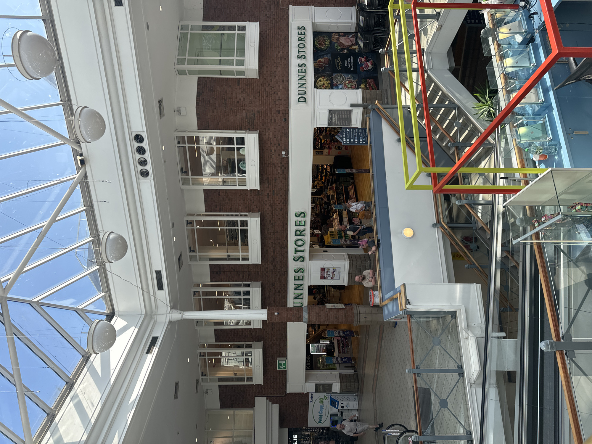 The image shows the interior of a shopping center, likely a Dunnes Stores. The architecture features a large glass ceiling, exposed brick, and modern escalators and railings. In the foreground, colorful metal frames (yellow and red) surround a display area with what appear to be electronic devices in glass cases. In the midground, people are visible inside the Dunnes Stores, browsing and shopping. A kiosk and a print shop are also visible. The Dunnes Stores signage is prominent, indicating it's a key establishment in the location. The presence of a sign with "Waterford City Square" suggests the shopping center is located in Waterford, Ireland. The overall scene depicts a bustling commercial environment on a bright, possibly sunny day, as indicated by the light filtering through the glass ceiling.