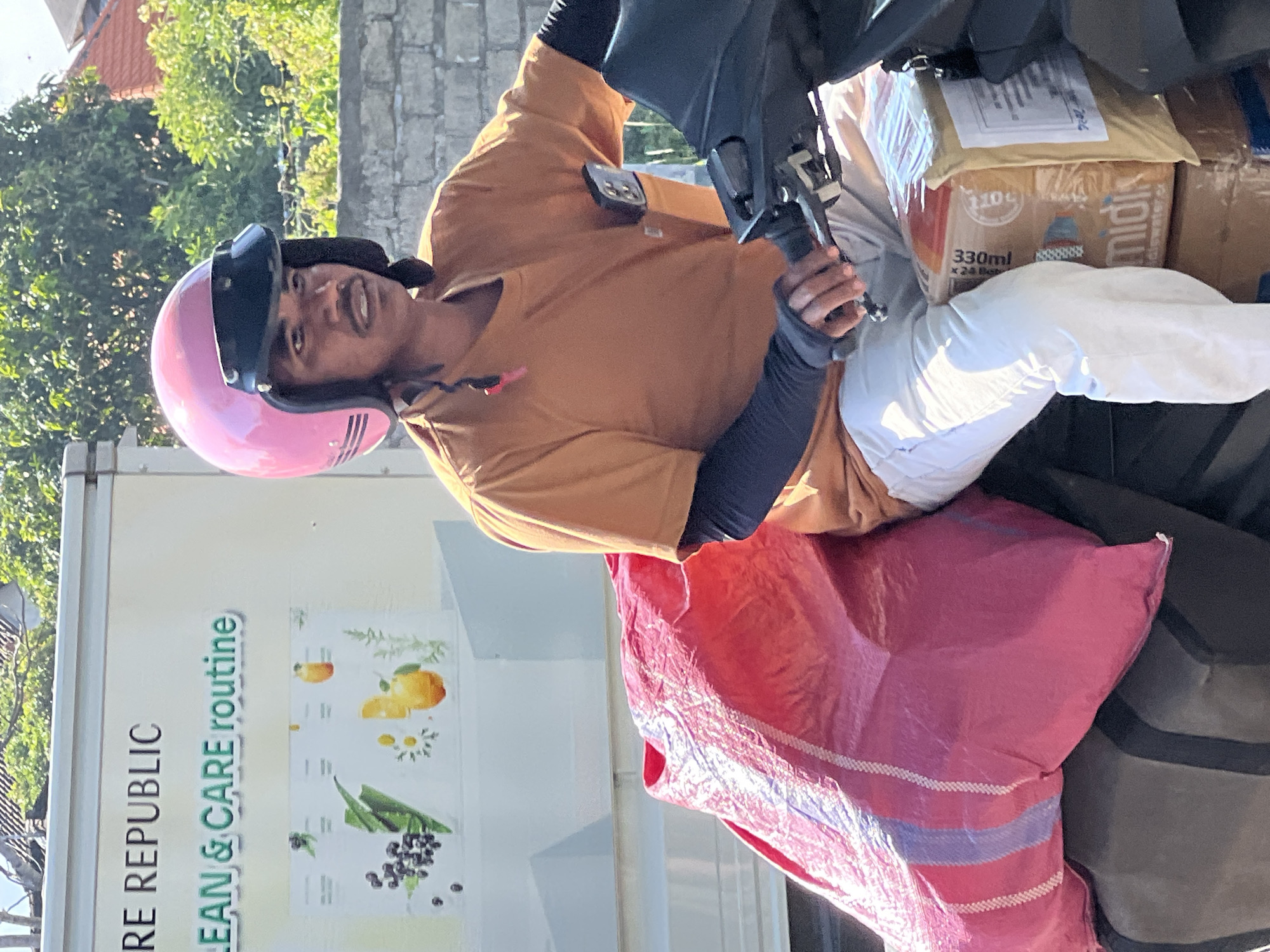 A man wearing a pink helmet and an orange t-shirt is riding a scooter loaded with packages. The scooter is in motion, with the man's hands on the handlebars. He is wearing white pants and black arm sleeves. A large pink bag is strapped to the back of the scooter, along with several cardboard boxes. The man appears to be a delivery driver, possibly in Denpasar, Indonesia, given the context provided by the prompt. The background shows a wall and lush green trees, indicating an outdoor setting. The man's facial expression is neutral.