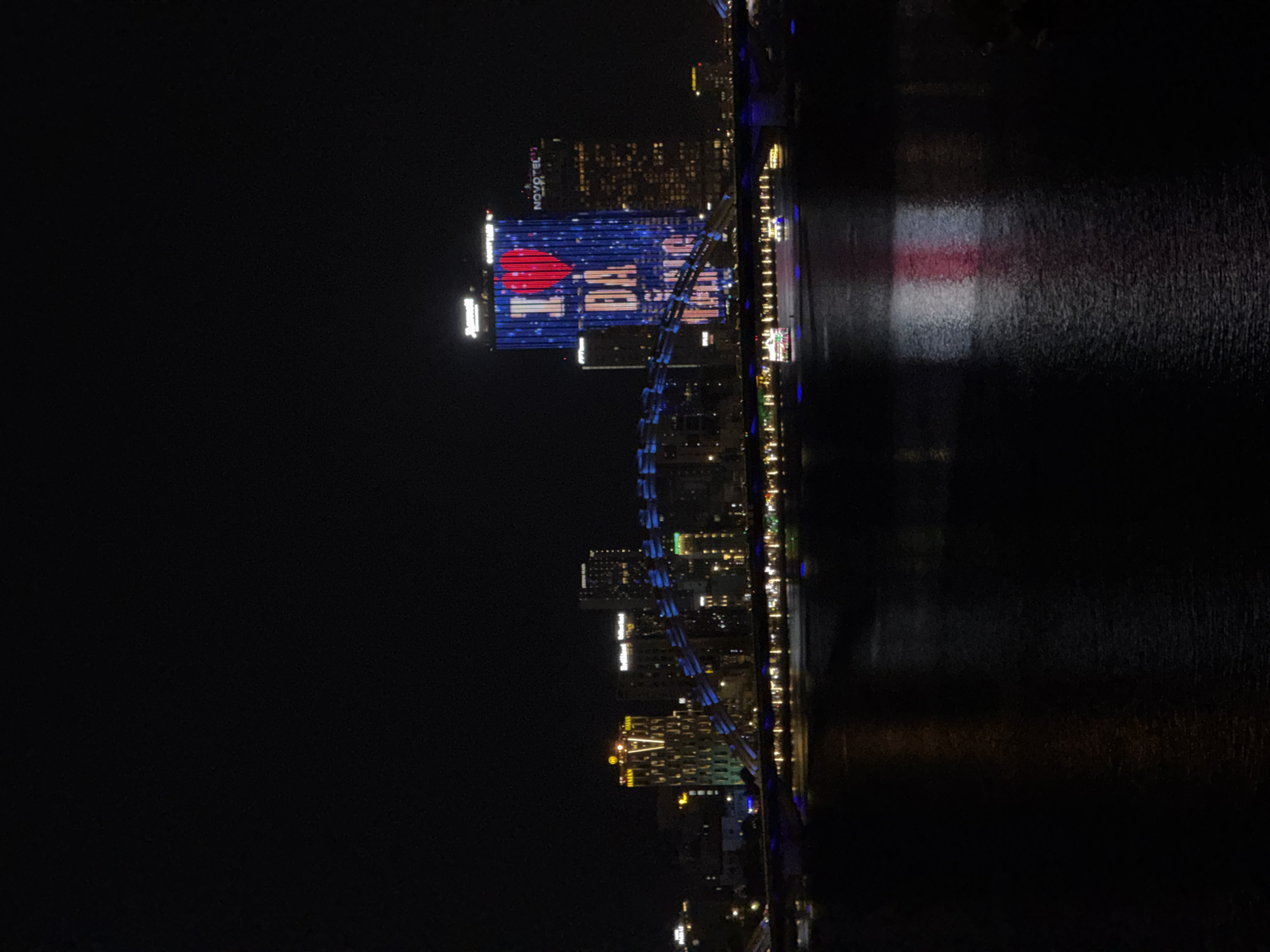 The image presents a stunning night-time view of a vibrant cityscape along a body of water, identified as Đà Nẵng, Vietnam. The scene is dominated by brightly lit buildings, their glow reflecting off the dark, rippled surface of what appears to be a river or ocean.
In the foreground, the dark water extends across the lower and right portions of the frame (when correctly oriented), capturing reflections of the illuminated structures. Notably, a distinct red and white reflection stretches across the water, mirroring the prominent light display on one of the buildings.
The main subject is a tall building on the city's riverbank, which features an elaborate light show. This display prominently showcases a large red heart symbol followed by the text "DA NANG", conveying the message "I ♥ DA NANG". Above this vibrant message, the hotel chain "NOVOTEL" is clearly visible in illuminated letters. Other multi-story buildings line the riverbank, their windows and facades lit up, contributing to the urban glow. Further along the bank, another building displays the distinctive "H" logo of the Hilton hotel chain. A decorative bridge-like structure, illuminated with blue lights, sweeps across the scene in front of the main buildings, adding architectural interest.
The time of day is undeniably night, with the sky appearing completely dark. The weather seems clear, allowing for crisp visibility of the city lights and their reflections. No people are discernible, and the activity captured is the static, impressive display of an illuminated modern city at night. The visible text, "I ♥ DA NANG", acts as a clear location cue and expresses civic pride.