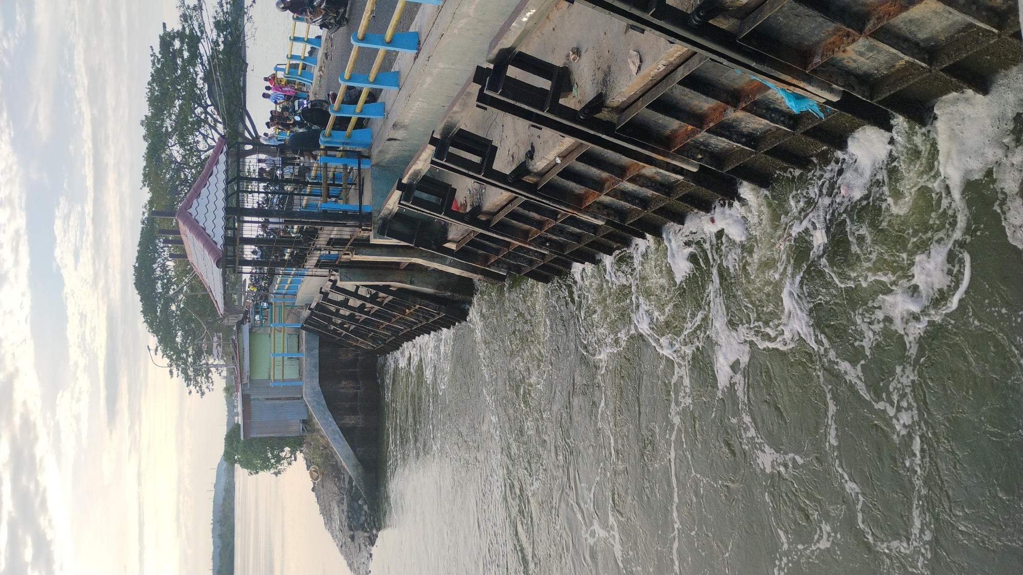 The image depicts the Krueng Cunda river dam in Lhokseumawe, Indonesia. The dam's concrete structure is visible, with metal gates allowing water to flow through, creating foamy ripples. The water appears murky green. On the dam's walkway, there is a small open-air pavilion with a tiled roof. Several motorcycles are parked under and around the pavilion. A group of people, some casually dressed and others in traditional attire, are gathered on the walkway, some talking, others observing the water. A blue and yellow railing lines the edge of the walkway. In the background, trees and a hazy sky with scattered clouds are visible. The scene suggests a public area where people gather, possibly for recreation or as a thoroughfare.